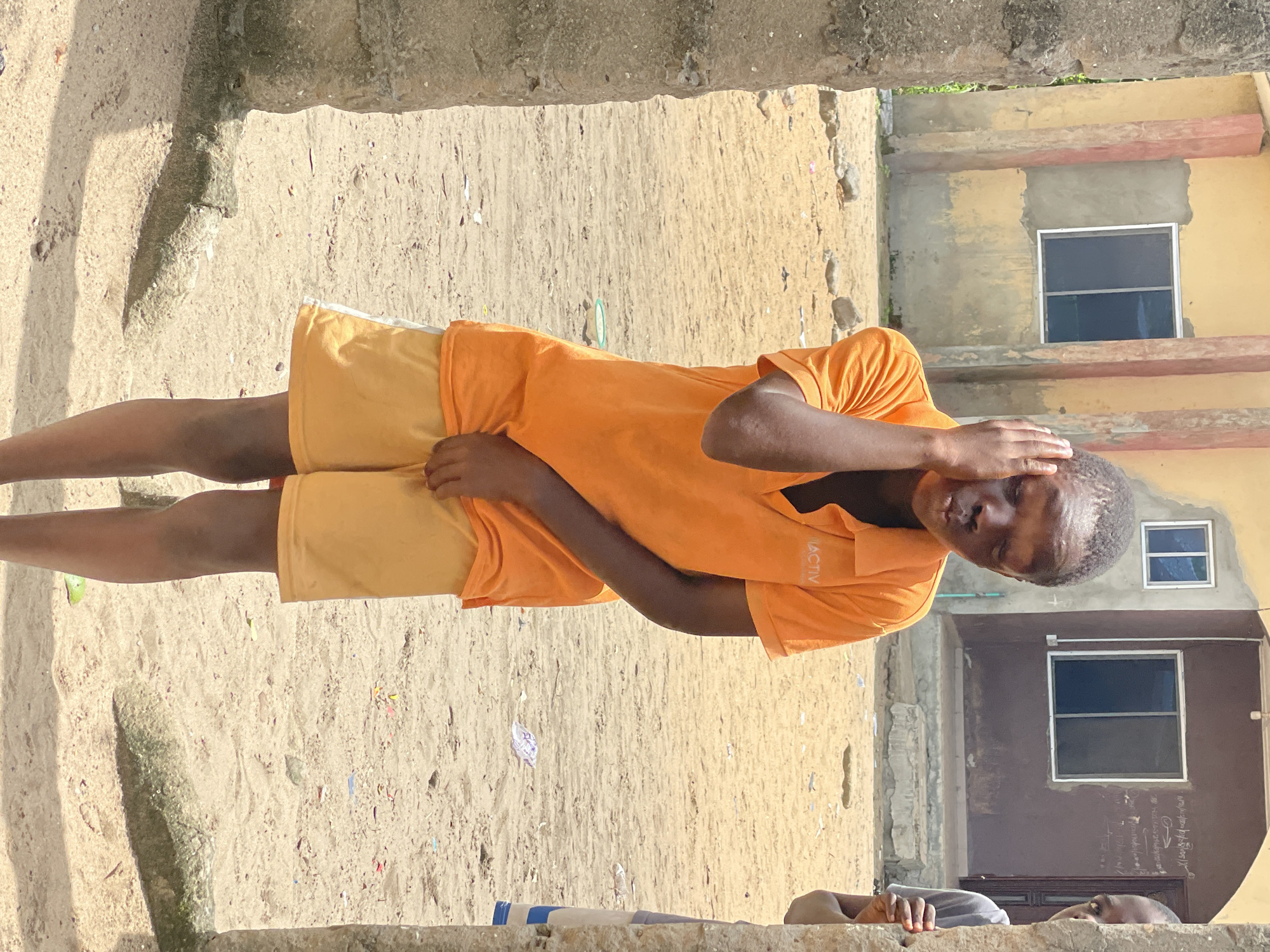 The image, taken in Badagry, Nigeria, shows a young person standing outdoors in bright daylight. The main subject, positioned slightly left of center, is a person with dark skin, wearing an orange short-sleeved t-shirt and yellow shorts. Their left hand is placed on their head, and they are looking towards the viewer with a somewhat neutral expression. The letters "ACTIV" are visible on the front of their orange t-shirt.
The setting appears to be an unpaved, sandy or dirt ground area. In the background, there are concrete buildings with simple architecture. One building, towards the right, is painted yellow and features a rectangular window with dark panes. Another building behind it, to the right, has a darker, possibly brown or gray, facade with a larger window and what looks like a door at the bottom. There are also visible concrete structures or walls framing parts of the scene, with some exposed rebar or rough edges.
A second person is partially visible in the bottom right corner of the frame, appearing to be behind a concrete ledge or wall. Only their head and shoulders are visible, and they are wearing a grey shirt. The ground is littered with small pieces of debris, including bits of colorful paper or plastic. The bright lighting suggests it is daytime.