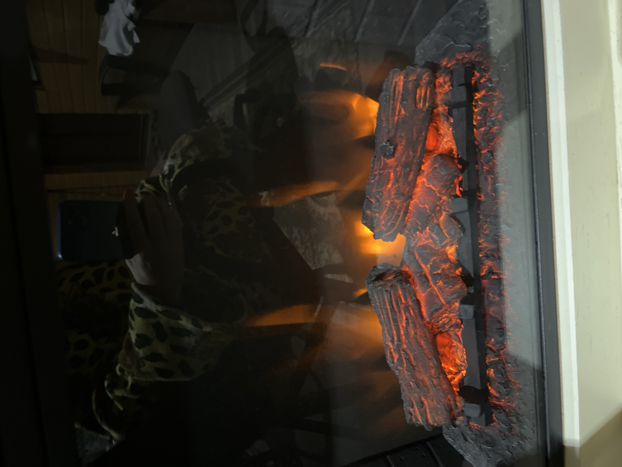 The image shows an electric fireplace with realistic-looking logs and glowing embers, viewed through glass. In the reflection on the glass, a person wearing a leopard-print robe is visible, holding a smartphone. The background of the reflection appears to be an indoor or covered outdoor space with wooden walls and furniture. There is no indication of the location being in the city of Saratov, Russia.