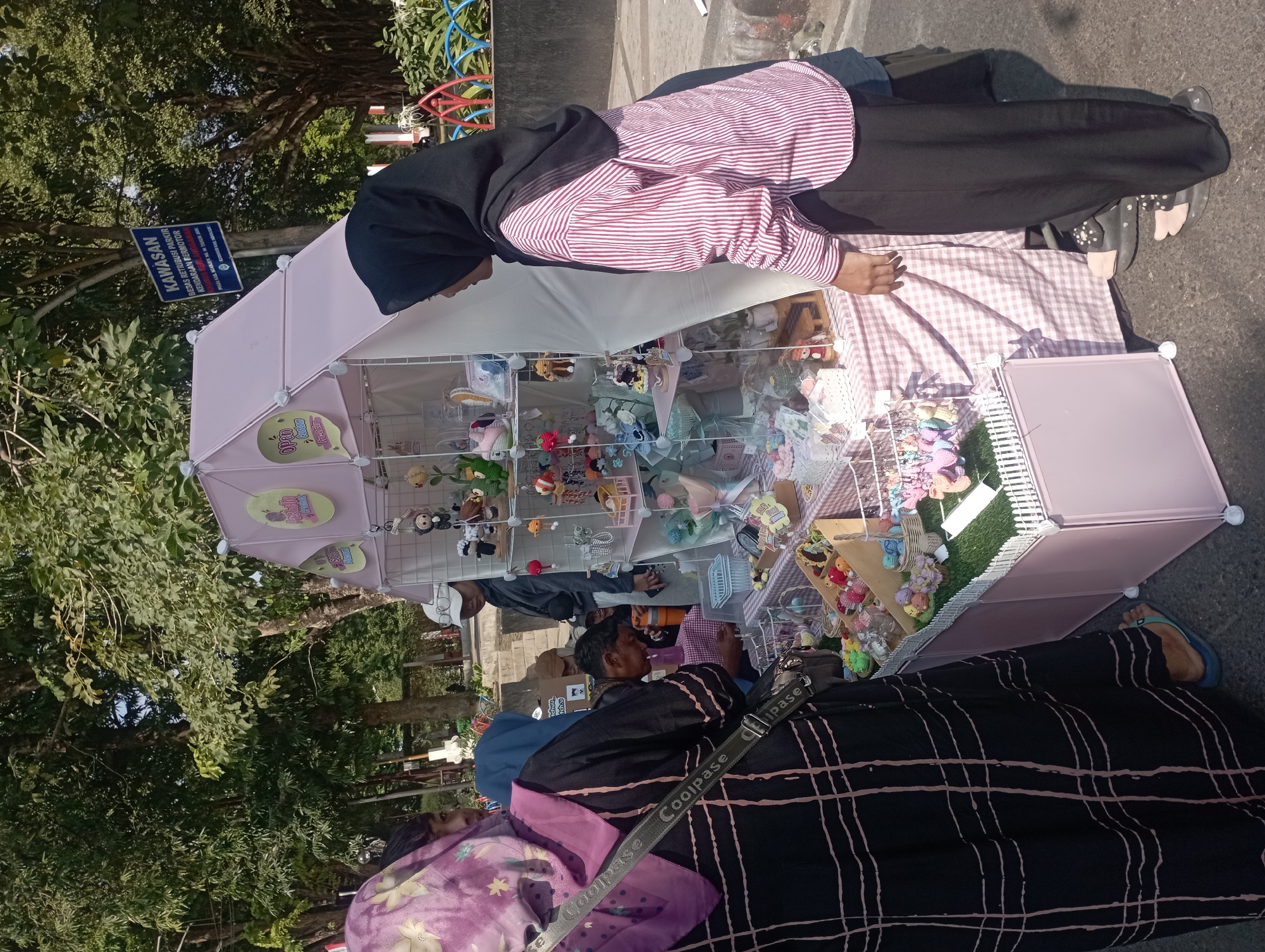 This image depicts a street scene in Nganjuk, Indonesia, likely during daytime given the strong sunlight and shadows. The focal point is a market stall selling handmade crafts, possibly keychains or small toys, displayed on tiered shelves within a pink, tent-like structure. The stall is covered with a pink and white checkered fabric. Several individuals are present, some appearing to be customers and others possibly vendors.
On the left, a person wearing a black garment with a purple and beige plaid hijab is looking towards the stall. To their right, another person in a blue hijab is also observing the merchandise. A man wearing a white cap and a grey jacket is seen behind the stall, interacting with the display. In the foreground and to the right, a person wearing a black hijab and a pink and white striped long-sleeved shirt is examining the items.
The environment surrounding the stall includes trees providing shade, a sidewalk or paved area, and some colorful playground equipment or decorative fencing in the background. A blue sign with white text, indicating "KAWASAN BEBAS RETRIBUSI PARKIR KENDARAAN BERMOTOR" (Motor Vehicle Parking Fee Exemption Area), is visible on a pole behind the stall. The overall impression is a vibrant outdoor market or fair.