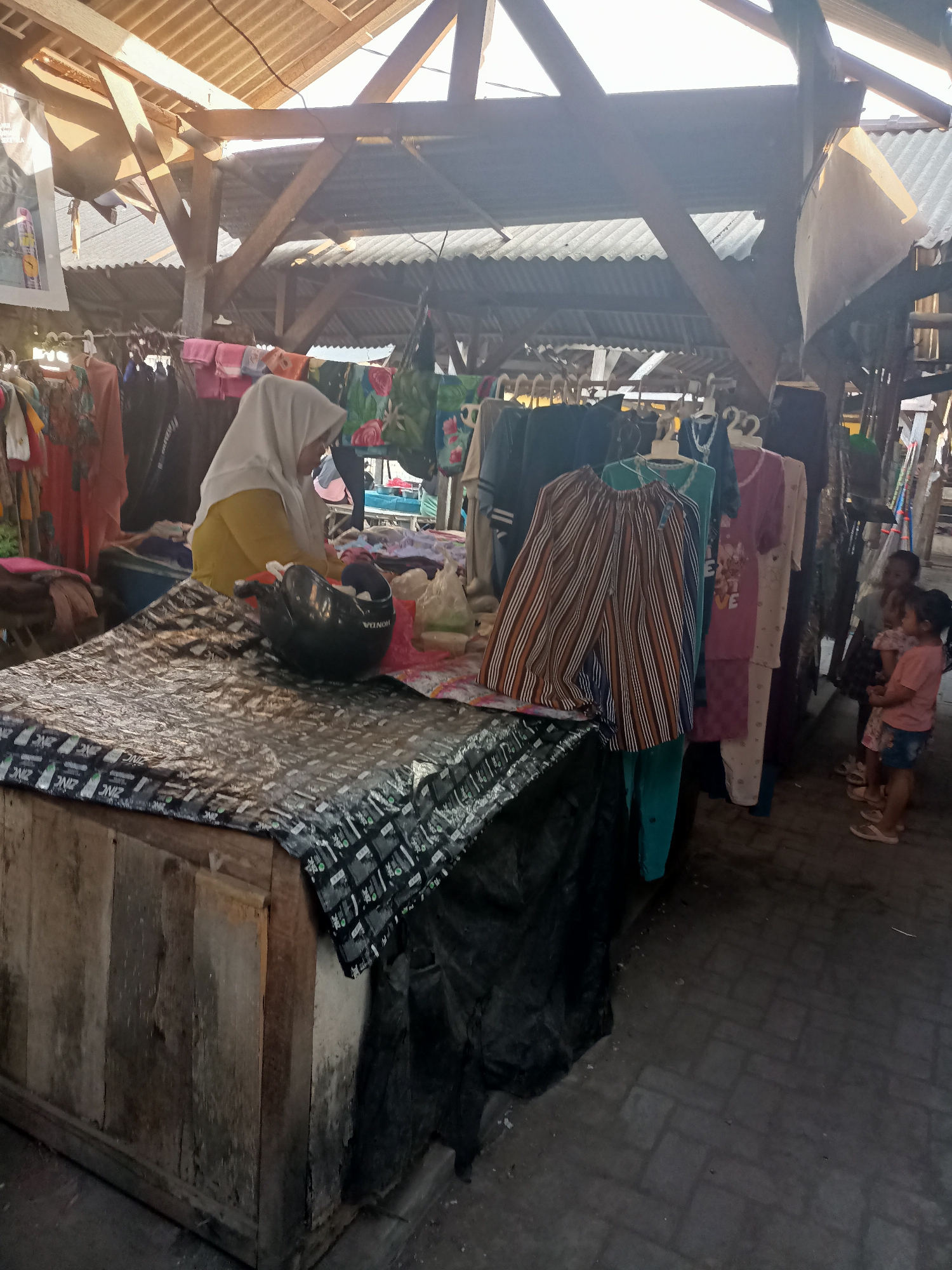 In the city of Badas, Indonesia, this image captures an outdoor market scene. A person wearing a yellow shirt and a white hijab is visible behind a wooden stall covered with a black tarp and a patterned sheet. On the stall, a black helmet rests near some items. Racks of clothing hang in the background, with various garments on display. Several children are also present in the area, with two younger girls in pink shirts and one older girl in a pink and white dress standing near the clothing racks. The market appears to be set up under a rustic wooden roof structure with corrugated metal sheeting. The ground is paved with interlocking bricks.