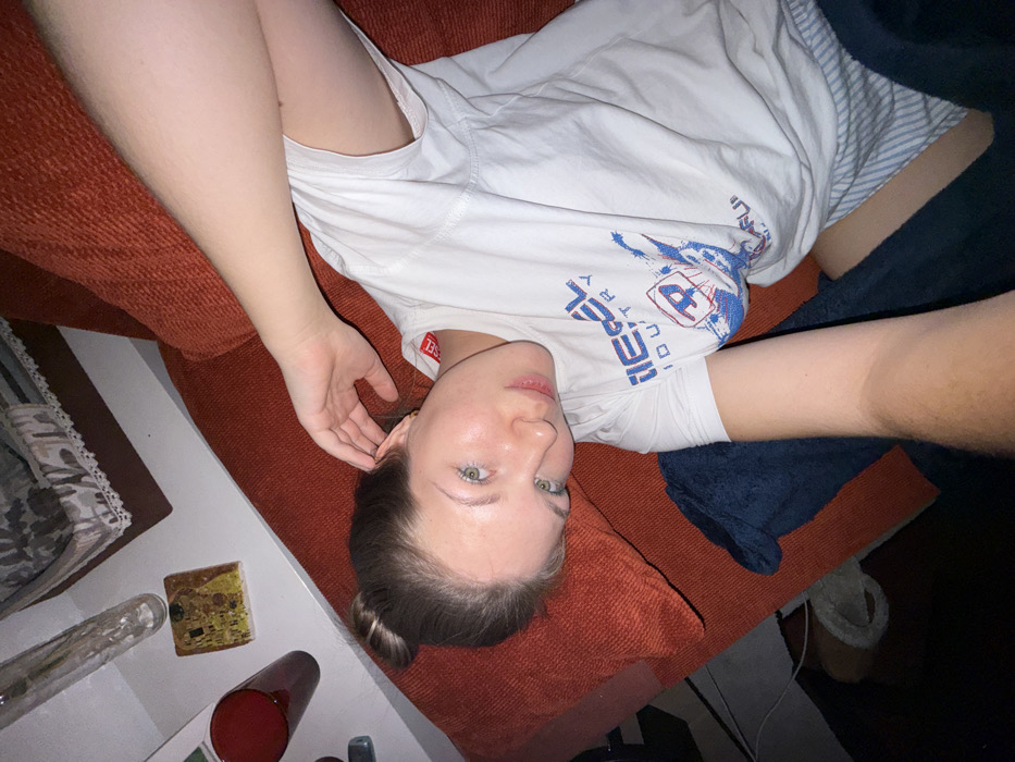 A woman lies on an orange couch. She is fair-skinned with light eyes, and her hair is pulled back in a bun. She is wearing a white t-shirt with a blue graphic. A portion of her legs, clad in striped underwear, are visible beneath a dark blue blanket. A side table next to the couch holds various items: a decorative square object, a red cup, and a long, clear object.