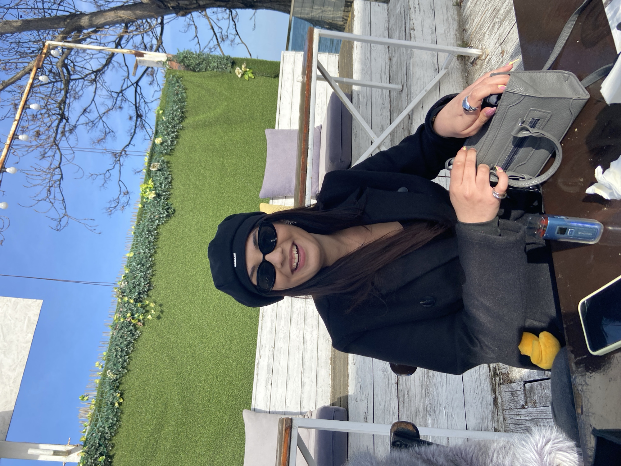 The image captures a woman sitting at a table outdoors. She is wearing a black beret and sunglasses and is dressed in a dark coat. The woman is smiling and looking towards the camera. She is holding a gray handbag in her hands and appears to be opening it.
The setting is an outdoor area with a white wooden fence in the background. A green wall of artificial plants and flowers provides a backdrop. The table is dark, and there are a few items on it, including what appears to be a cellphone and a small yellow object. There are cushions on the seats, and bare branches of trees are visible in the upper left corner against a blue sky. The location of the city is Unknown, Unknown.