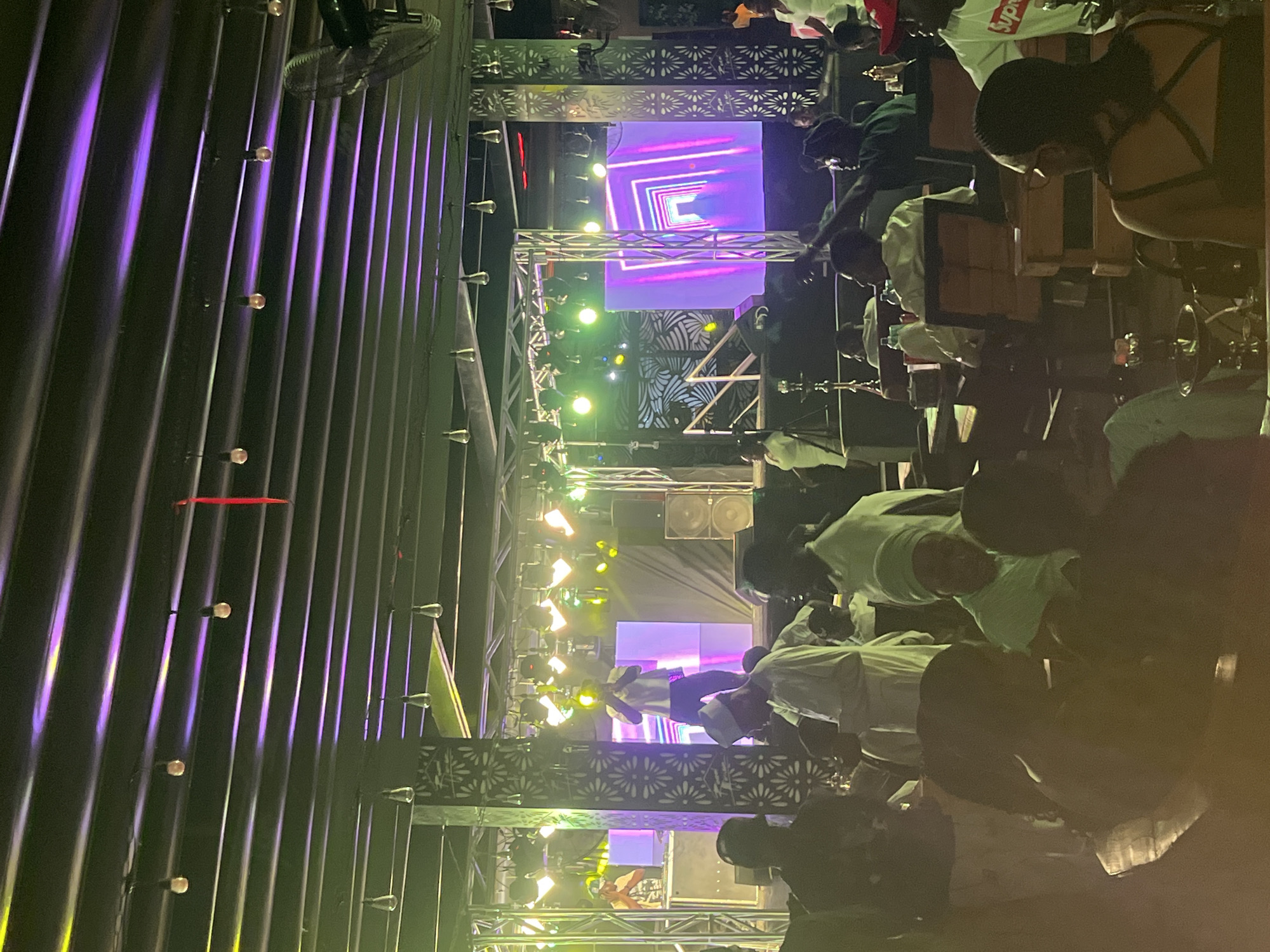 The image depicts a crowded indoor event, likely a nightclub or concert venue, in Howayi, Nigeria. The scene is brightly lit with stage lighting and illuminated vertical corrugated metal walls. A stage structure with patterned metalwork supports lighting fixtures and a large video screen displaying geometric patterns. Numerous people are present, mostly in the foreground, observing the stage. Most are dressed casually. Seated individuals are visible at tables with drinks. The overall atmosphere suggests a lively and crowded social gathering.