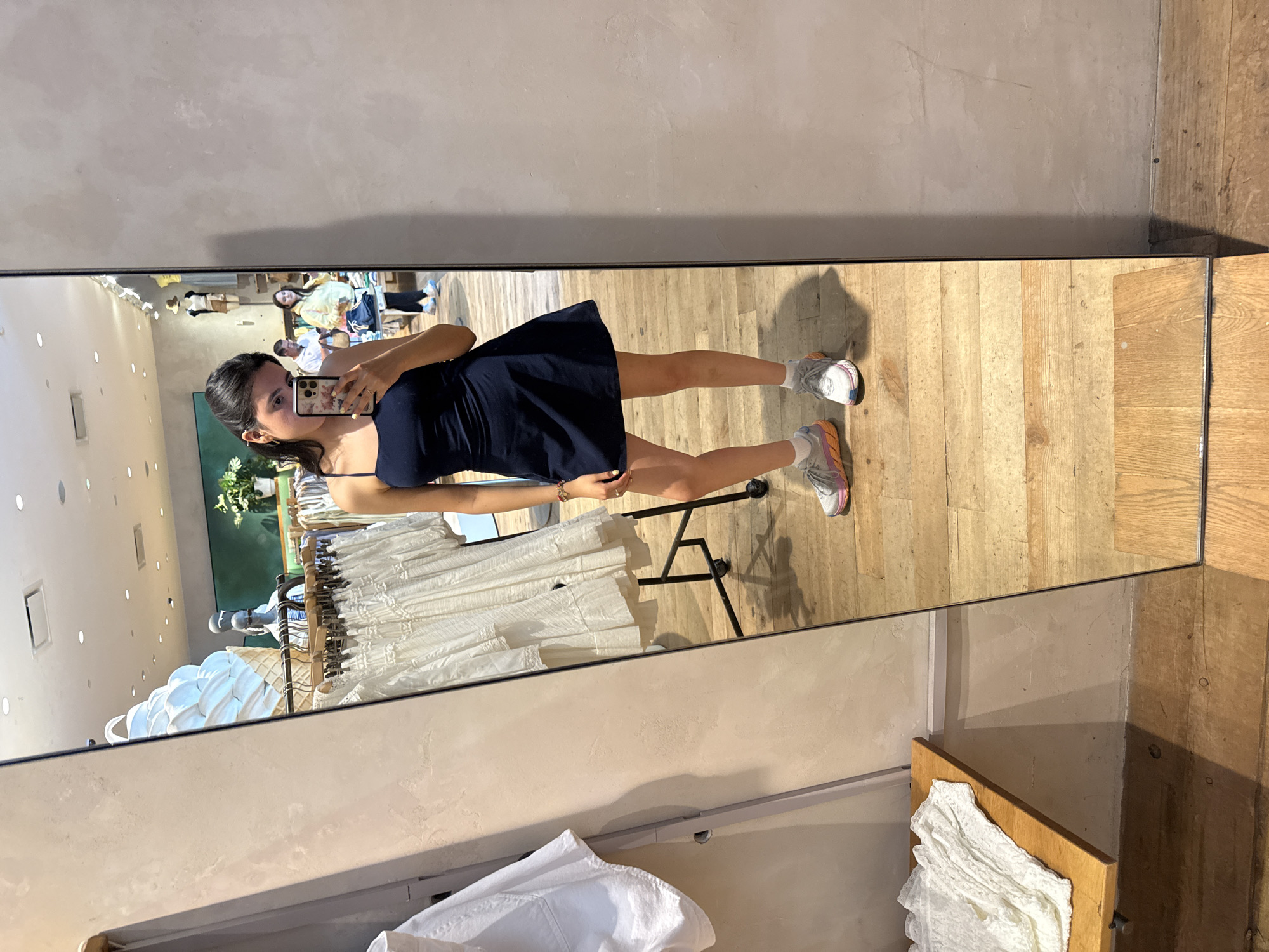 A young woman stands in front of a large mirror inside a retail store, presumably trying on clothing. She is holding a smartphone and taking a photo of herself in the mirror. She is wearing a dark-colored dress and sneakers with light-colored socks. The mirror reflects the interior of the store, including racks of clothing and other shoppers.
The store appears to be located in Austin, Texas, United States of America.