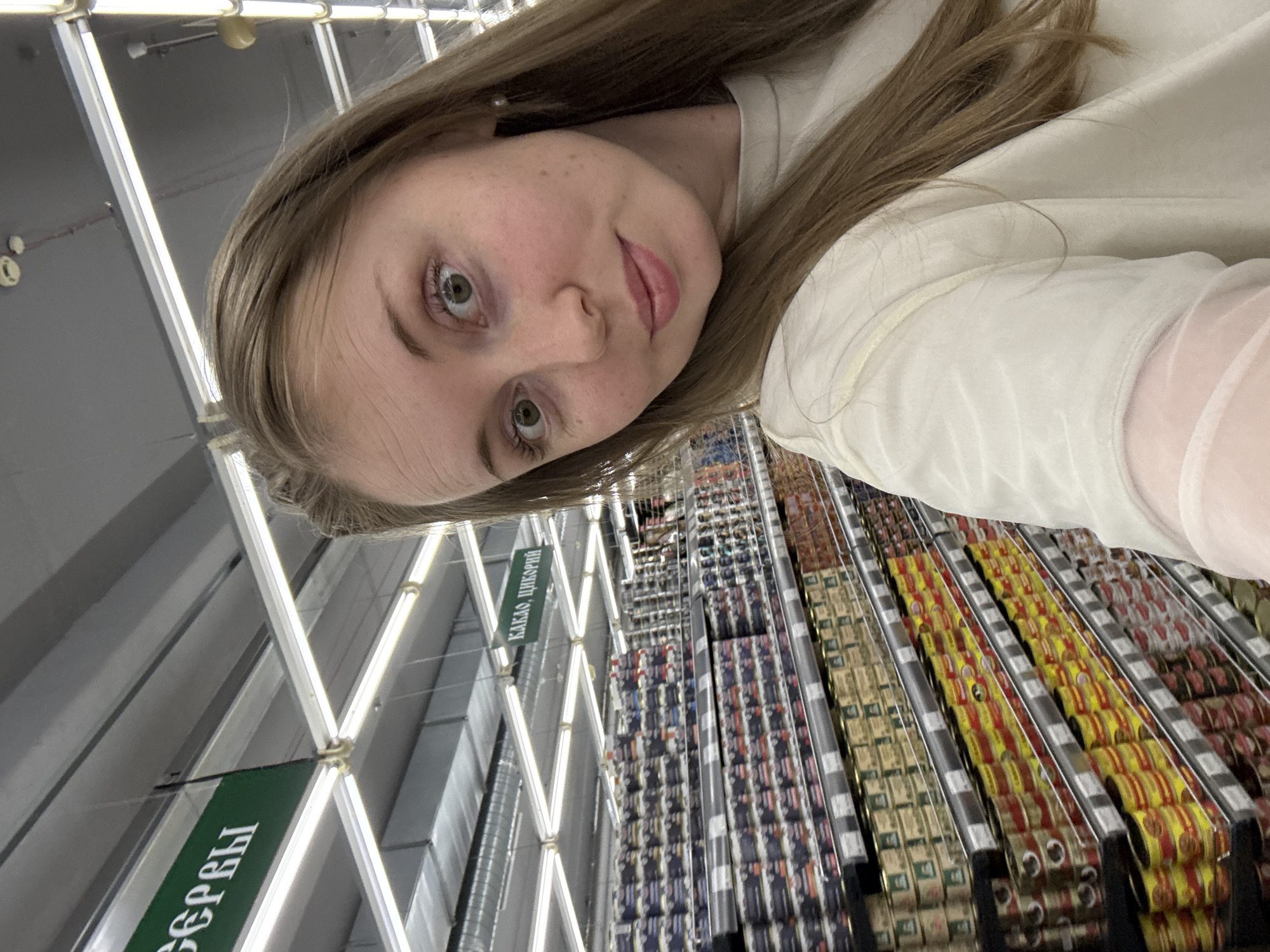 A young woman is captured in a selfie within a supermarket aisle. She possesses light brown hair, green eyes, and fair skin, looking directly into the camera with wide eyes and a subtle smile. She is wearing a light-colored, possibly off-white, top with a sheer sleeve detail visible on her right arm. The background features extensive shelving units stocked with numerous canned goods of various colors, including dark blue, yellow, and red. Overhead, a grid of fluorescent lights illuminates the aisle, alongside visible ventilation ducts and ceiling-mounted sensors. A green sign hanging from the ceiling displays Cyrillic text, reading "КАКАО, ЦИКОРИЙ" (Cacao, Chicory), while another partial sign shows "СЕРЫ".