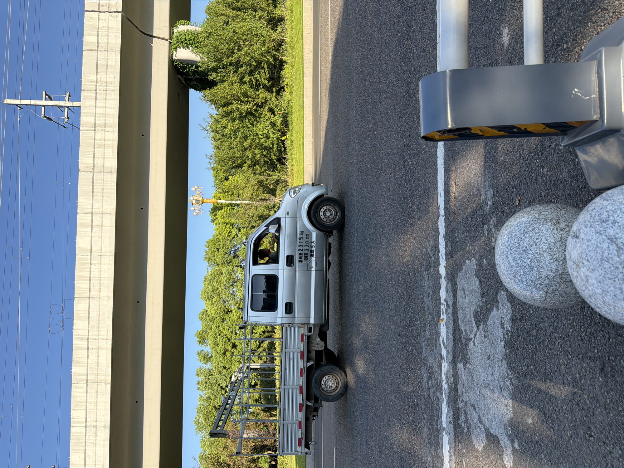 A silver flatbed truck with a metal cage on its bed is driving on an asphalt road. The truck has Chinese text on its side, including "总质量 2315 KG" (total mass 2315 kg), "额定载荷 370 MM" (rated load 370 mm), and "准乘 5人" (passenger capacity 5 people). A person is visible driving the truck. In the foreground, there are two large, spherical decorative stones and a section of a traffic barrier with yellow and black diagonal stripes. In the background, there is a concrete overpass, trees, and power lines against a clear blue sky. The scene appears to be in Jiangjin, China, as indicated by the text on the truck and the urban infrastructure. The lighting suggests it is daytime, likely late afternoon due to the shadows.