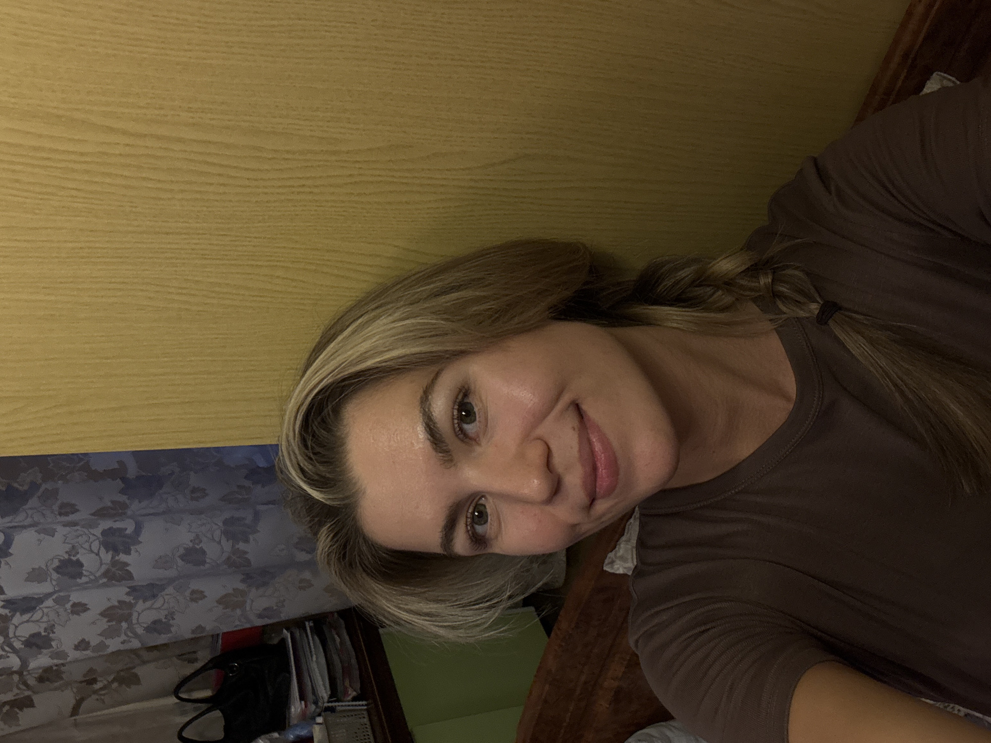 A woman with light brown and blonde hair, styled with a side part and a braid secured with a black tie over her left shoulder, is the primary subject of the image. She has light-colored eyes and a gentle smile, looking directly into the camera. She appears to be wearing a plain, short-sleeved brown top.
The setting appears to be indoors. In the background, to the upper left of the frame, there is a light-colored wooden surface, possibly a wall or a headboard, displaying prominent wood grain. Below this wooden surface, on the lower left, a patterned fabric with blue and brown leaf designs, likely a curtain or sheet, is visible. Further to the bottom left, some dark objects, resembling a bag and stacked papers or books on a shelf, are partially seen. On the right side of the frame, a darker wooden furniture piece is present.
The woman's head is tilted significantly to her right, indicating the photograph was taken at an angle, possibly a selfie. The lighting is consistent with an indoor environment, potentially with artificial light, creating a warm and somewhat subdued atmosphere. There is no visible text in the image.