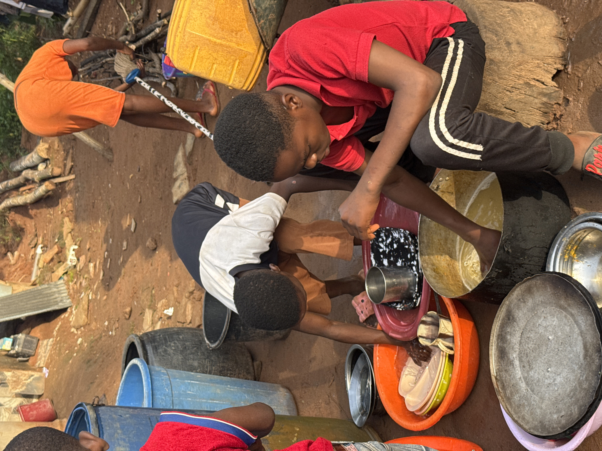 In an outdoor scene, likely a rural or semi-urban setting, at least three young individuals are engaged in a communal activity under bright daylight. The ground is a reddish-brown dirt, with scattered logs, branches, and discarded corrugated metal sheets visible.
Two individuals are kneeling and washing dishes. The person on the right, wearing a red t-shirt and black track pants with white stripes, is actively scrubbing a metal pot in a yellow basin filled with soapy water. To their left, another individual in a black and white t-shirt is also washing dishes in an orange basin, which contains several plates and bowls. Both have their hands submerged in the water. Various other metal pots, pans, and silver cups are arranged around them, some clean, some waiting to be washed.
Further back and to the left, another person in an orange t-shirt and shorts is standing, holding a mop with a black and white patterned handle. This individual appears to be either starting to clean or has just finished. A large yellow plastic jerrycan is situated near them. Numerous plastic buckets and basins of different colors (blue, black, red, orange) are visible throughout the scene, some stacked or used for holding water or items.
The overall atmosphere suggests a chore being carried out, possibly after a meal, with the individuals focused on their tasks. On the sole of a black sandal worn by the person in red and black, the word "DOPE" is visible in red text. The bright lighting indicates a clear, sunny day.