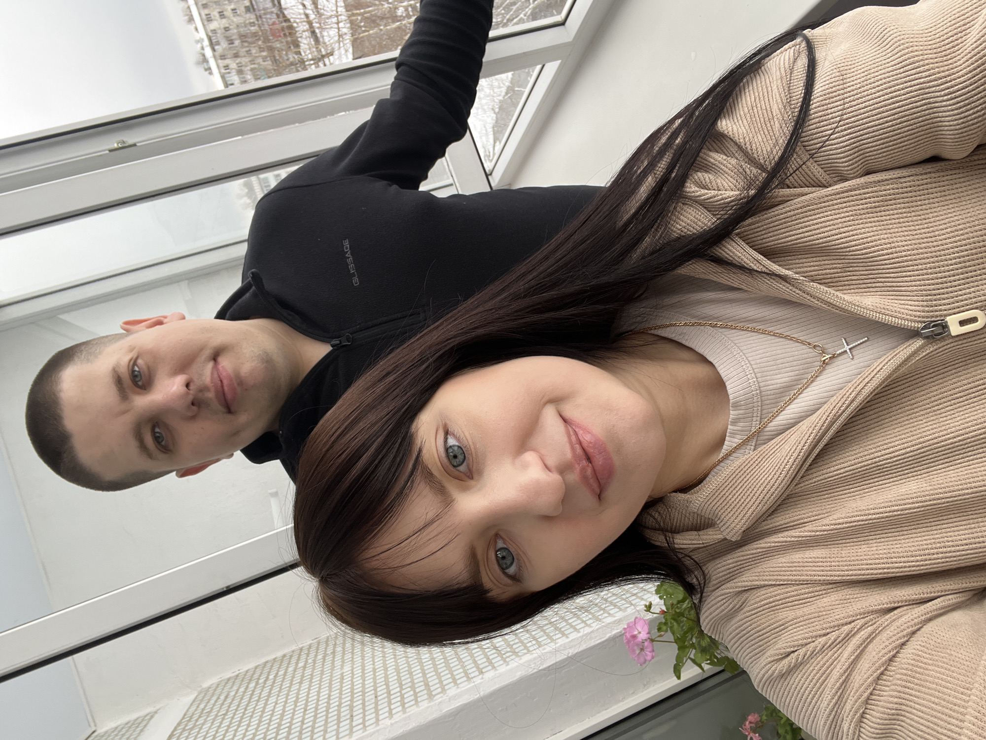 Two individuals, a male and a female, are in close proximity indoors, likely taking a selfie. The male, with short, cropped hair, is wearing a black zip-up sweatshirt with the text "GLISSADE" visible on the left chest. The female has long, dark hair and is wearing a beige ribbed zip-up top over a lighter-colored shirt, with a delicate gold necklace featuring a cross pendant. The background features a window with a view of a multi-story building and leafless trees, possibly indicating winter. The location is likely the city of Chaikovsky, Russia. The individuals appear to be interacting by posing for the photo. Pink flowers are visible near the lower right corner of the image. The building visible through the window appears to be an apartment building.
