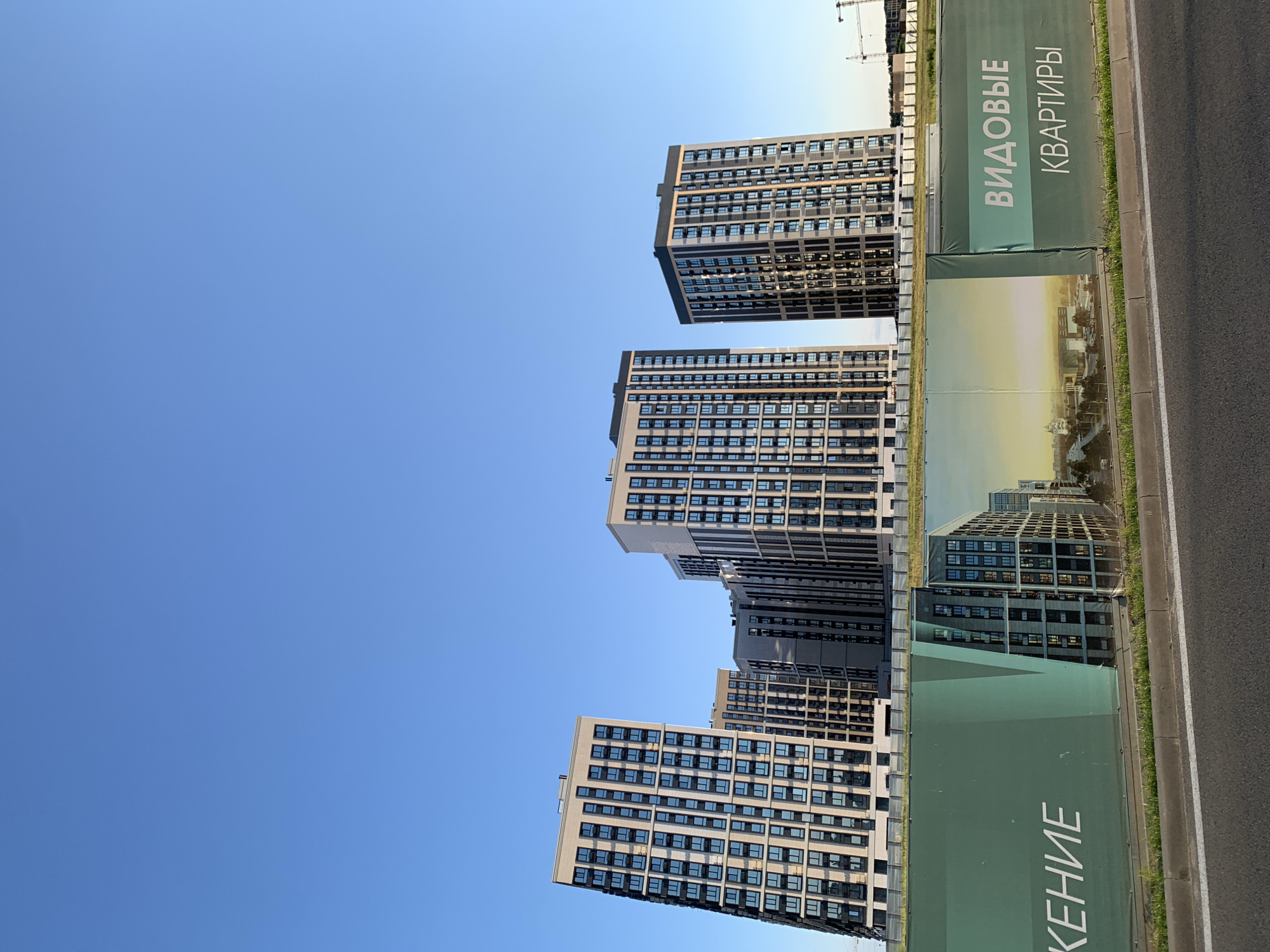 The image shows a construction site in Stavropol, Russia, with several modern apartment buildings under construction. A large green banner spans the foreground, displaying text in Russian, which appears to be an advertisement for apartments. The banner features an artist's rendering of a city skyline and some of the apartment buildings. The sky is clear and blue. There are no people visible in the image. The overall impression is one of urban development and new construction.
