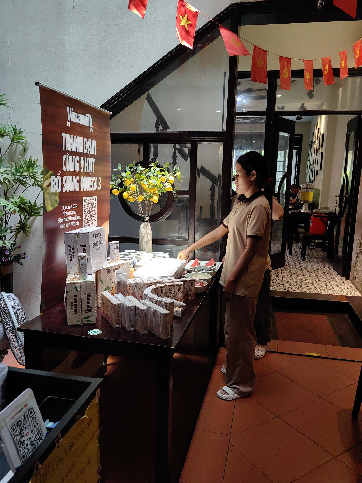 An indoor scene in Hà Nội, Vietnam, captures a Vinamilk product promotion or tasting event. In the foreground, a young woman wearing glasses, a beige polo shirt, and light brown pants is reaching towards a display table laden with Vinamilk products and snacks. Behind her, another person's lower legs are partially visible.
The central feature is a dark brown display table holding various Vinamilk cartons and boxes, primarily "Sữa hạt 9 loại hạt" (9-nut milk) and "Sữa Yến Mạch" (Oat Milk). Several bowls of cut fruit, including watermelon slices, are also present, alongside a decorative vase with artificial yellow citrus fruits and green leaves. To the left of the table, a white electric fan is visible.
A prominent banner stands behind the table, featuring the "Vinamilk EST 1976" logo and text in Vietnamese: "THANH ĐẠM CÙNG 9 HẠT BỔ SUNG OMEGA 3" (Lightly sweet with 9 nuts, supplemented with Omega 3). Below this, a QR code and instructions read: "Quét mã QR Nhận ngay Sữa hạt 9 loại hạt Sữa Yến Mạch" (Scan QR code to get 9-nut milk and Oat Milk immediately). Another QR code is visible in a container on the floor, possibly for payment or information.
The setting appears to be a cafe or restaurant, characterized by dark wooden door frames and a patterned tiled floor. Overhead, a string of red and yellow Vietnamese flag bunting, featuring both the star and hammer-and-sickle symbols, adds a festive nationalistic touch. In the background, through an open doorway, more of the interior is visible, including a large beverage dispenser with a yellow liquid and a red bucket on a patterned floor. The general ambiance suggests an organized event promoting dairy and plant-based milk products.