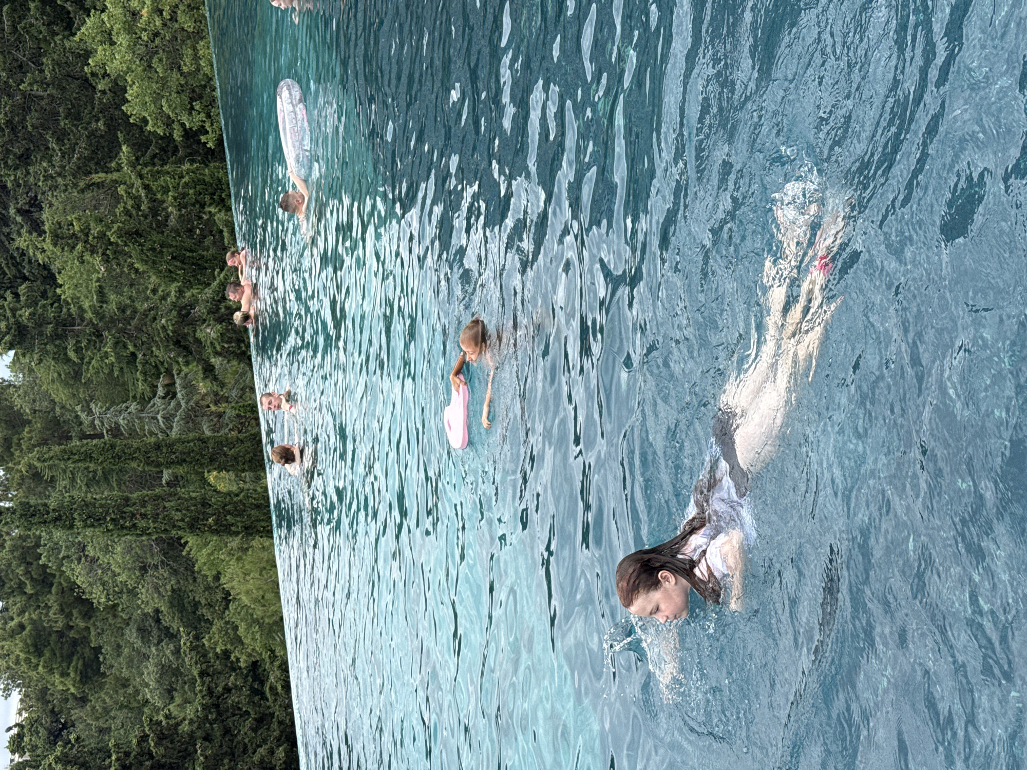 An aerial view shows an infinity pool at the edge of a lush green forest with a clear blue sky. Several people are scattered throughout the pool, some swimming, some relaxing, and others conversing. In the foreground, a young girl with long brown hair is swimming towards the left, her face tilted upwards. She is wearing a white t-shirt and dark shorts. In the middle ground, two children are playing in the water. One child, a girl with blonde hair, is reaching for a pink inflatable. Another child, a boy, is behind her. Further in the background, a group of adults are socializing at the edge of the pool. The water is clear and inviting, and the surrounding trees create a serene and natural backdrop. The overall impression is one of leisure and relaxation in a beautiful outdoor setting.