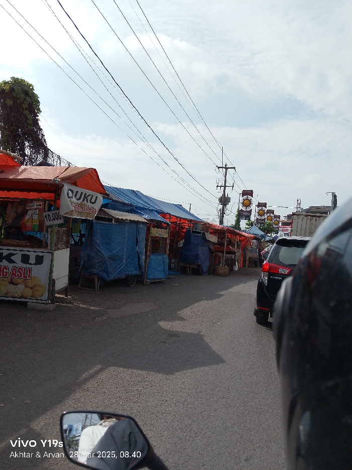The image captures a roadside market scene in Jatibarang, Indonesia, at 08:40 AM on March 28, 2025, as indicated by the timestamp. The perspective is from a vehicle, likely a motorcycle, with a dark helmet partially visible in the foreground on the right and a rearview mirror reflecting a person in a white shirt at the bottom left.
Along the left side of a paved road, numerous makeshift stalls are set up, featuring simple frames covered by brightly colored tarpaulins, primarily orange and blue. Some stalls appear open for business, while others are entirely covered with blue tarps, suggesting they are closed or protecting goods. One sign on a prominent stall on the left clearly displays "DUKU" (a type of fruit), "Rp 10.000" (a price in Indonesian Rupiah), and "ASLI" (authentic).
The day is bright and sunny, with a partly cloudy sky. Numerous overhead electrical wires and utility poles stretch across the frame into the distance. Further down the road, red and yellow banners featuring sun-like logos are strung across the street. A black car, possibly an SUV, is visible on the right side of the road, behind some stalls. Shadows are distinctly cast on the road and ground, consistent with morning sunlight. The scene depicts a typical busy local marketplace or street, with vendors prepared for daily commerce.