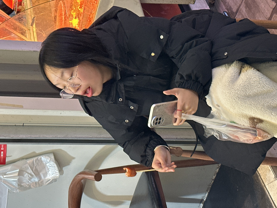 The image depicts a young East Asian female seated in what appears to be a casual food establishment in Jiangjin, China. She has straight black hair and wears round, wire-rimmed glasses. She is dressed in a black puffer jacket and has a cream-colored fleece garment or blanket on her lap. Her mouth is slightly open, revealing her tongue, and she is looking upwards and to her right.
In her left hand, she holds a light-colored smartphone with a multi-lens camera array visible on its back. A transparent plastic bag containing what appears to be raw meat or another food item hangs from her left hand, below the phone. A thin red string or bracelet is visible on her left wrist. Her right hand holds a wooden skewer with a single piece of orange-brown, cooked food, possibly a fried sausage or similar item.
She is seated on a chair with a dark brown wooden frame and a black upholstered seat, partially visible on the left. The background consists of a light-colored wall with a clear plastic bag hanging from it on the left. To her right, a glass display case is visible, illuminated by a warm, reddish-orange light, showcasing items that appear to be food, consistent with a food stall or restaurant setting.