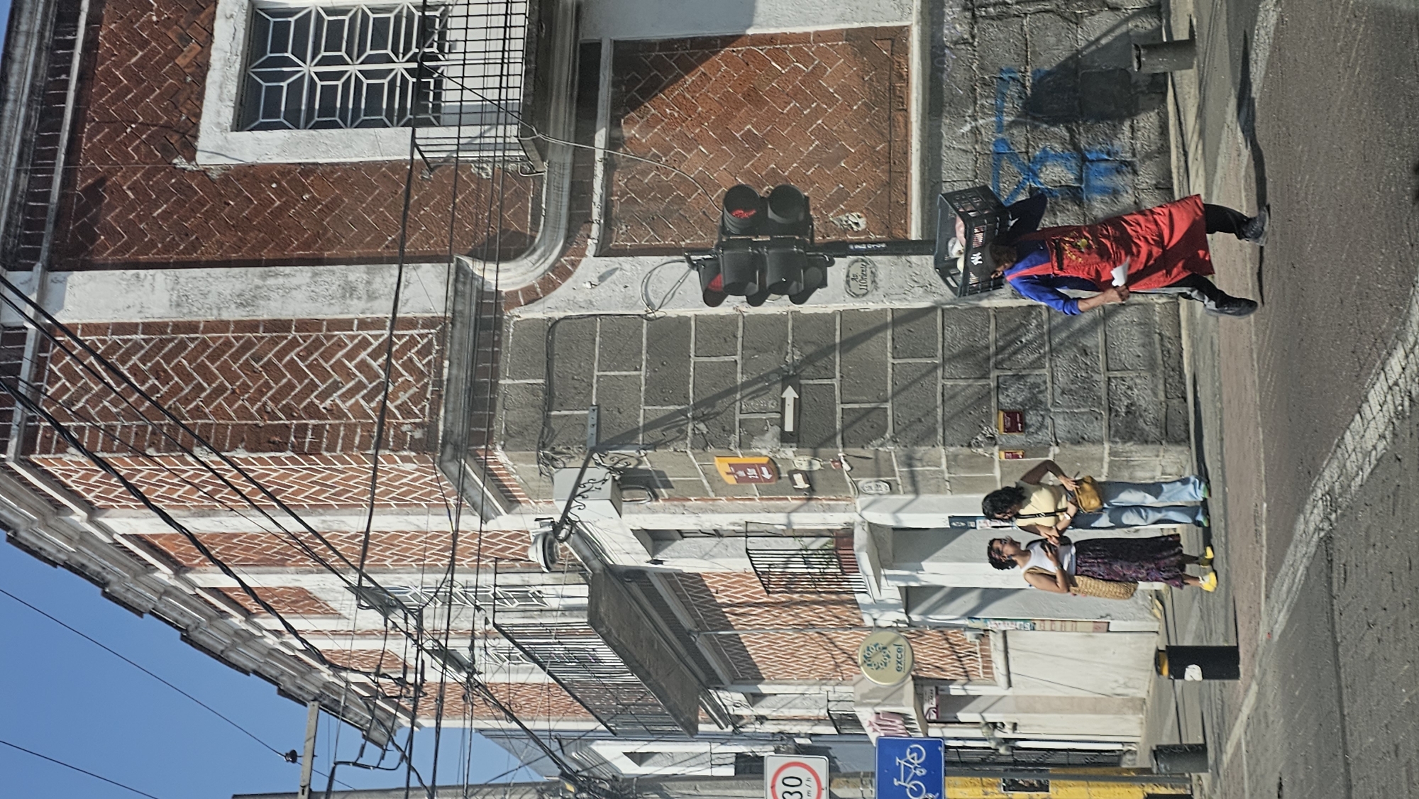 A street scene captured in bright daylight shows buildings, a sidewalk, and an asphalt road under a clear blue sky. The image is oriented vertically, with buildings on the left and the street on the right.
The multi-story buildings feature red brick facades with white mortar, arranged in distinct patterns, including herringbone. Windows are protected by decorative white metal grates, and some sections include balconies with black metal railings. Numerous black power lines extend across the frame, obscuring parts of the buildings and sky.
A grey tiled sidewalk runs along the buildings. A black traffic light, displaying a red signal, hangs over the street. Below it, a white arrow is painted on the road surface, pointing forward. Further down the street, a black bollard and a street lamp are visible. Blue spray-painted graffiti appears on a lower brick wall.
Three people are present. One individual, possibly a vendor, wears a red apron over a blue shirt and carries a black plastic crate on their back, walking along the edge of the street. Two other people stand on the sidewalk. One wears light blue jeans, a light yellow top, and a yellow bag around their waist. The other wears a patterned long skirt, a white sleeveless top, and bright yellow shoes, carrying a woven bag.
The asphalt street shows a white painted line. Notable visible text includes a red circular sign displaying "30 km/h" and a blue square sign with a white bicycle symbol. Part of a shop sign that reads "excel" is also visible. The strong shadows cast by the buildings and people indicate a sunny day.