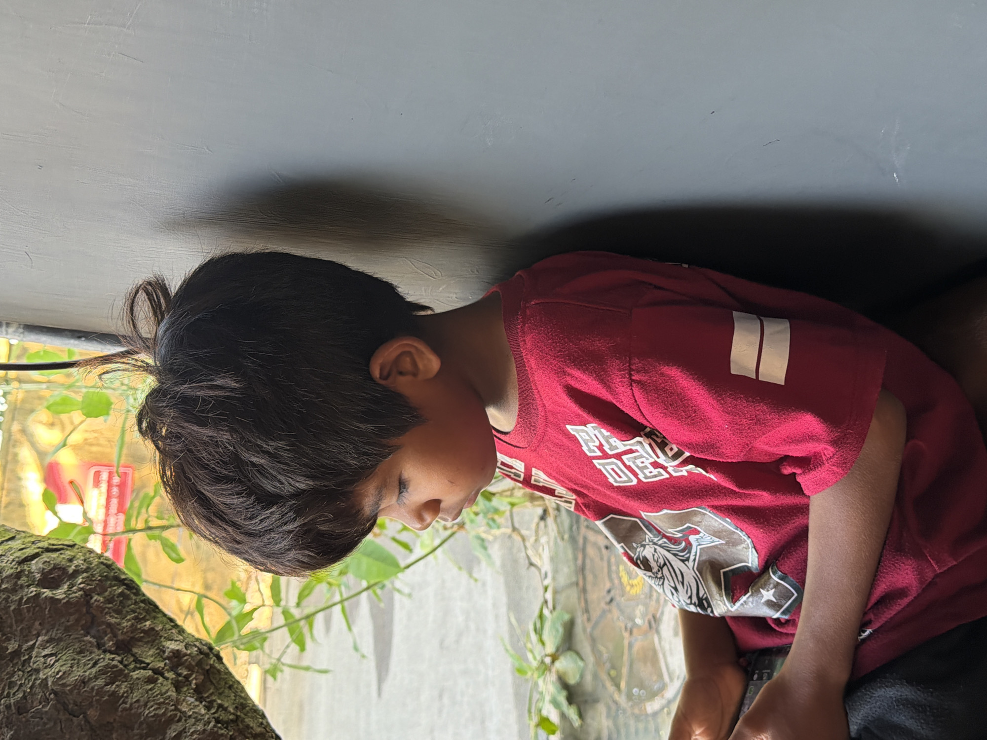 In a side-profile shot, a young boy with dark hair is captured from the chest up. He is wearing a red t-shirt with white detailing on the sleeves and a graphic design on the front. The boy is looking down and to his left, with his face partially obscured by his hair. He is leaning against a plain gray wall, with his left shoulder visible. To the boy's left, a large, textured tree trunk partially frames the scene. In the background, lush green foliage is visible, with a yellow wall and some indistinct red lettering behind it. The overall lighting suggests an outdoor environment, possibly in a semi-shaded area. The image was taken in Mengwi, Indonesia.