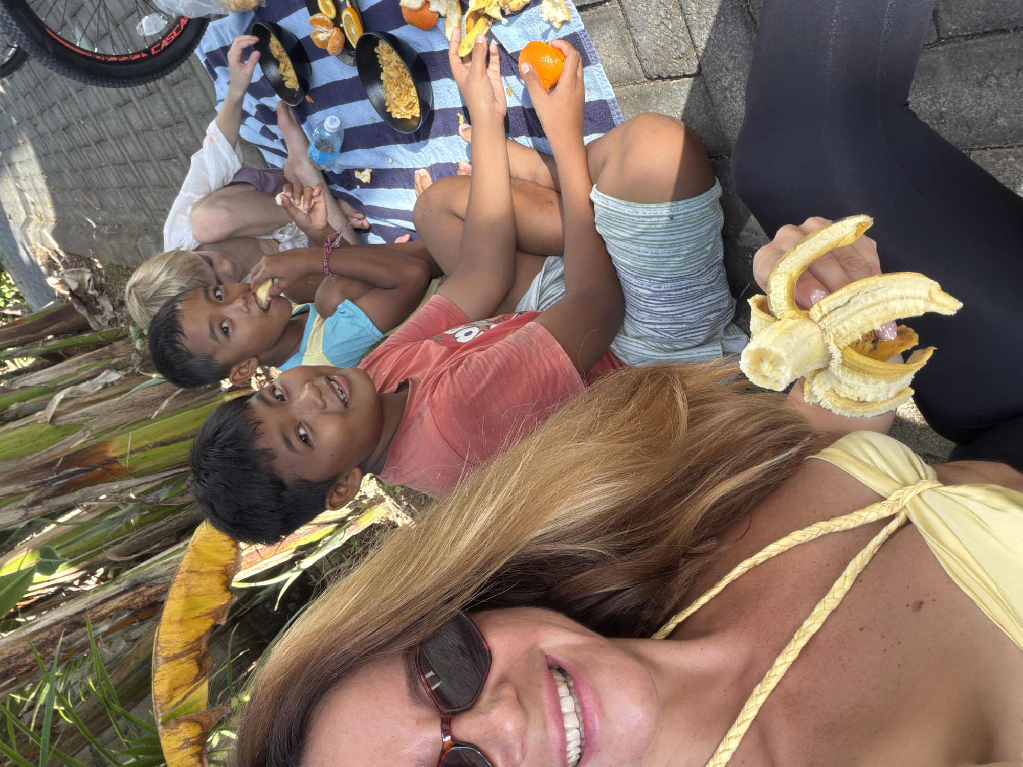 In Kuta Selatan, Indonesia, a woman with blonde hair and sunglasses smiles as she holds a peeled banana. Beside her, two boys and another person sit on a striped blanket on a paved area, surrounded by lush greenery and what appear to be banana trees. The group is enjoying snacks, with bowls of cereal and sliced oranges visible on the blanket. A bicycle wheel is also present in the background. The scene suggests a casual outdoor gathering.