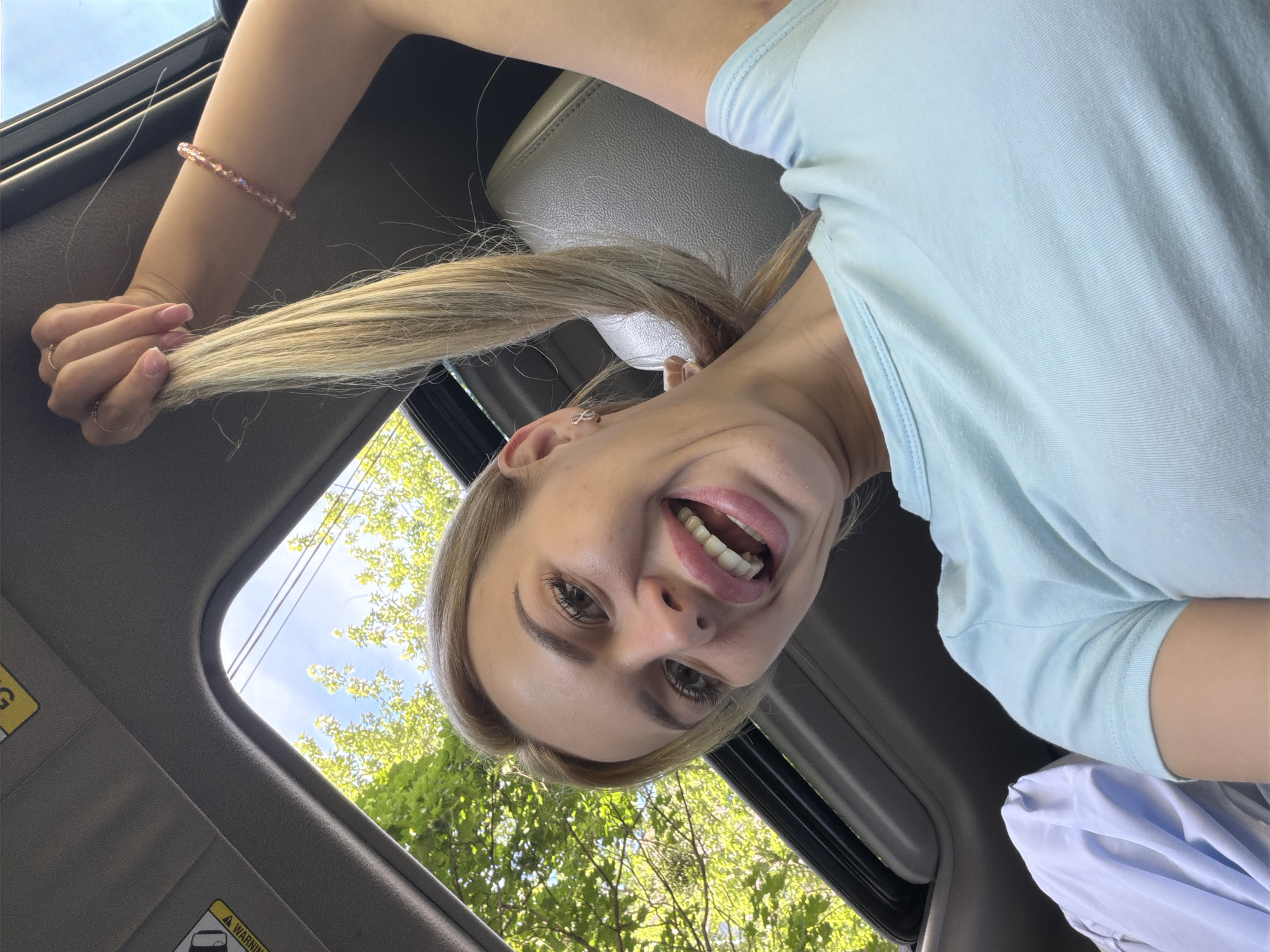 A young woman with blonde hair is in the passenger seat of a car, looking up at the camera. She is smiling broadly, with her mouth open, and her blonde hair is being pulled upwards. She is wearing a light blue t-shirt and a beaded bracelet on her right arm. The car's sunroof is open, revealing green trees and a blue sky. The background shows the interior of the car, including the grey leather seats and the dark grey trim around the sunroof. This image was taken in Kyiv, Ukraine.