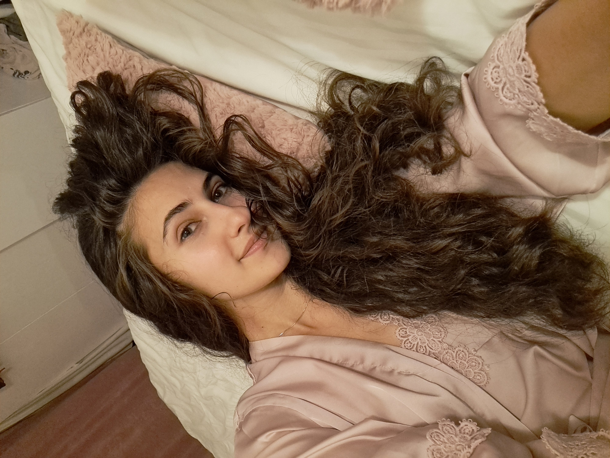 The image displays a young woman lying on a bed, looking towards the camera with a soft, slight smile. Her long, dark brown, voluminous curly hair is spread out around her head on the white bedding. She is wearing a light pink, silky or satin robe adorned with intricate lace trim details visible along the lapel and sleeves. A delicate gold chain necklace is also visible around her neck.
The setting appears to be an indoor space, likely a bedroom, with white sheets and a fluffy pink pillow positioned behind her head. A plain light-colored wall is seen in the background, along with a hint of a floor or carpet. The lighting is soft and ambient, consistent with an indoor environment, possibly in the evening or at night. There is no visible text in the image. The overall impression is one of comfort and tranquility.