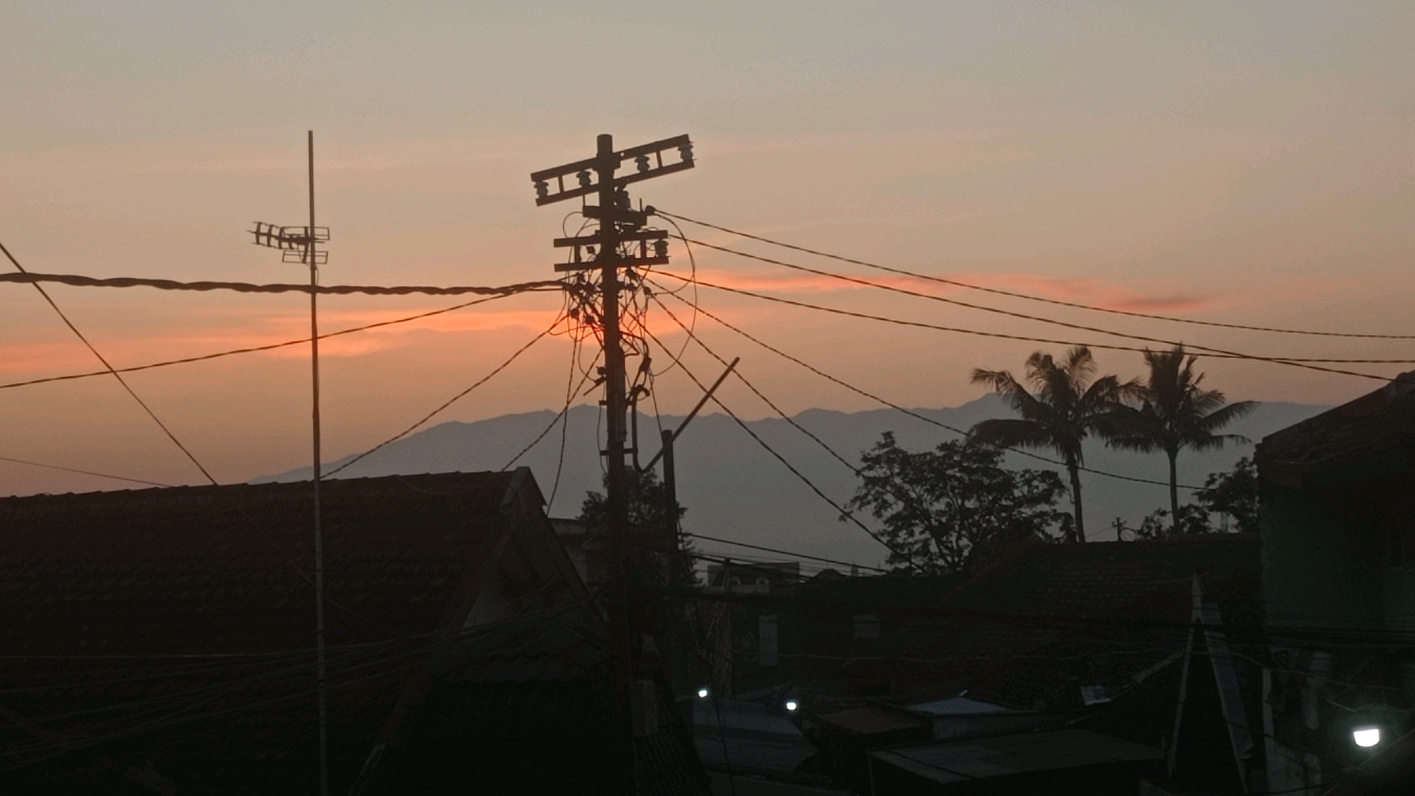 The image depicts a silhouetted landscape at dusk or dawn, likely in Mulyoagung, Indonesia. The sky is a gradient of pale orange and gray, with a faint strip of brighter orange clouds near the horizon. In the foreground, a dark, complex network of power lines crisscrosses the frame. A prominent utility pole, also silhouetted, stands to the right of center, heavily laden with wires and electrical equipment.
To the left, a smaller utility pole with an antenna is visible. In the midground, the dark silhouettes of rooftops are discernible, suggesting a dense residential or urban area. Beyond the rooftops, a line of indistinct mountains or hills forms the backdrop. Two palm trees stand silhouetted against the sky between the utility pole and the mountains. The overall impression is one of a typical, somewhat cluttered, urban or semi-urban environment at a transitional time of day. No people are visible in the image.