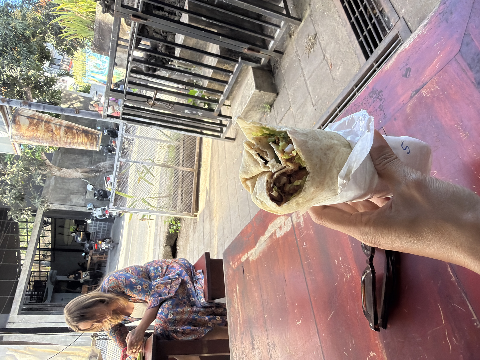 A person is holding a wrap in front of a red wooden table. On the table are a pair of folded sunglasses. In the background, a woman in a blue and orange dress is leaning over the table. Behind her is a building with open sides. Outside the building, there are motorbikes parked along the street. The photo was taken in the Kuta Selatan region of Indonesia.