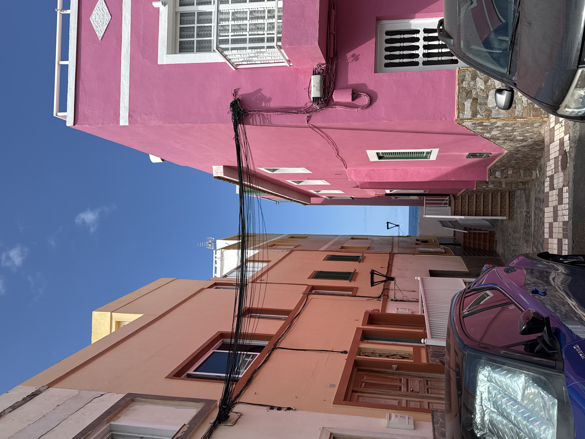 A narrow street is shown with two brightly colored buildings facing each other. The building on the left is painted in shades of peach and orange, with wooden doors and windows. The building on the right is painted a vibrant pink, with white trim and a balcony. The street is paved with cobblestones. The sky is blue with a few white clouds. In the distance, the ocean can be seen. Two cars are parked on the street, one is a purple hybrid car with a reflective sunshade in the windshield, and the other is a dark SUV. Electrical wires are strung across the street between the buildings.