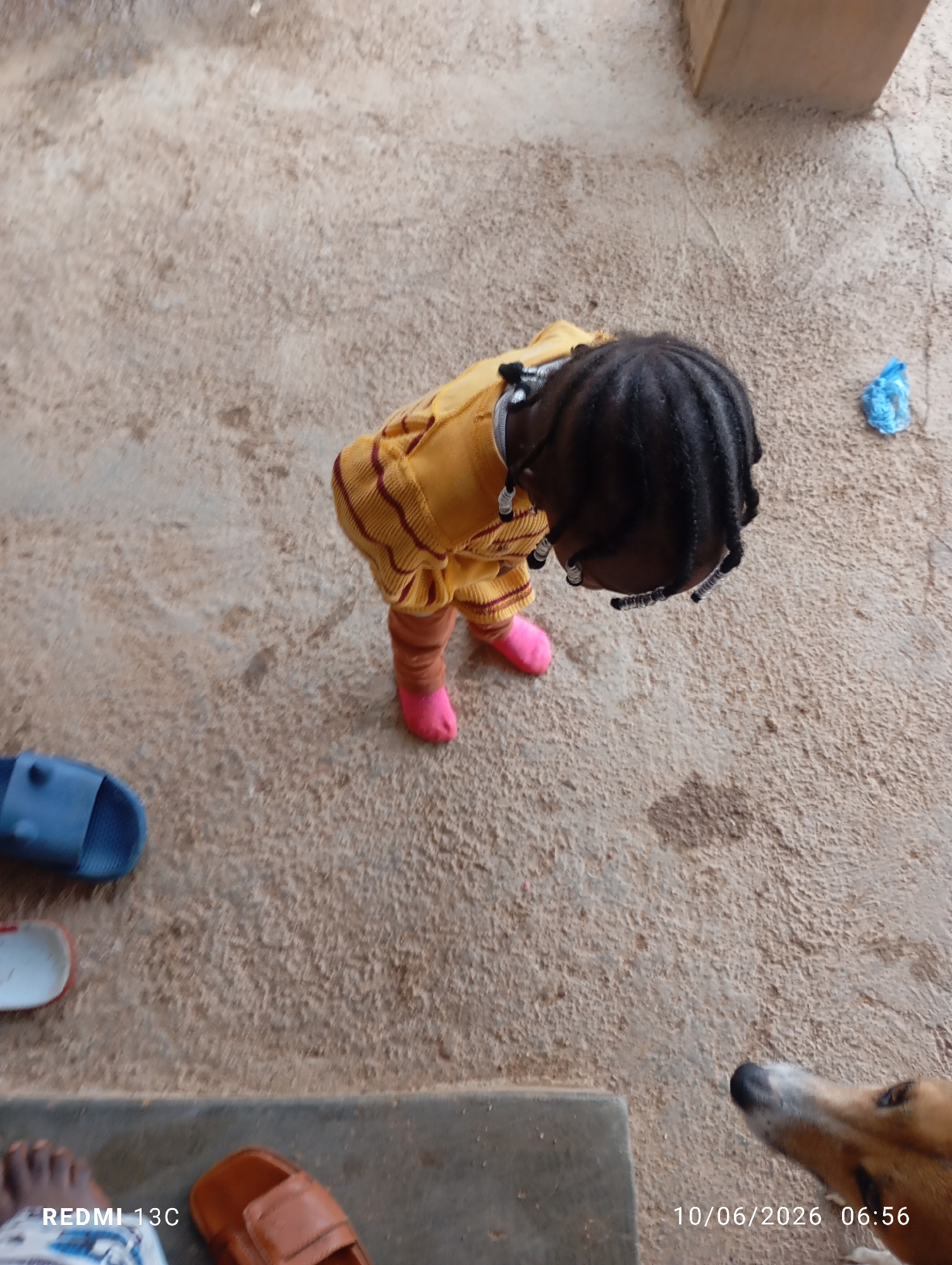An outdoor, overhead shot captures a young child wearing a yellow and striped garment and pink socks, standing on a sandy or concrete surface. Adjacent to the child is a blue sandal. Further to the left, another sandal, partially visible, rests on the ground. In the lower right corner, a dog's head is visible, looking upwards, and a timestamp reads "10/06/2026 06:56". The ground is uneven and appears to be made of a textured, light brown material, possibly dirt or concrete. A small, crumpled blue object is visible in the upper right portion of the image. The bottom left corner displays "REDMI 13C".