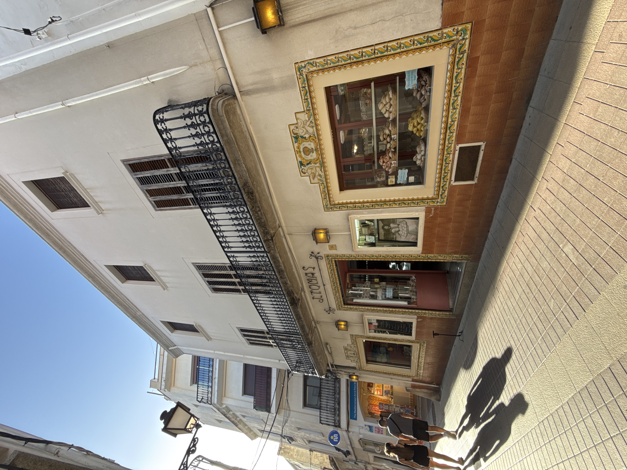 The image shows a narrow street in Tossa De Mar, Spain, bathed in sunlight. In the foreground, a couple walks away from the camera, their long shadows cast on the tiled pavement. The man wears a grey t-shirt and dark shorts, and the woman wears a black tank top and dark shorts. To the right, a white building with brown tiled lower facade features a bakery or pastry shop called "J. TOMAS" with a prominent display window showcasing various pastries. The building has a balcony with black wrought-iron railings and shutters on the upper windows. A street lamp is visible on the left side of the image. Further down the street, another building with a blue circular sign and a shop front selling art prints is visible. The sky is clear and blue.