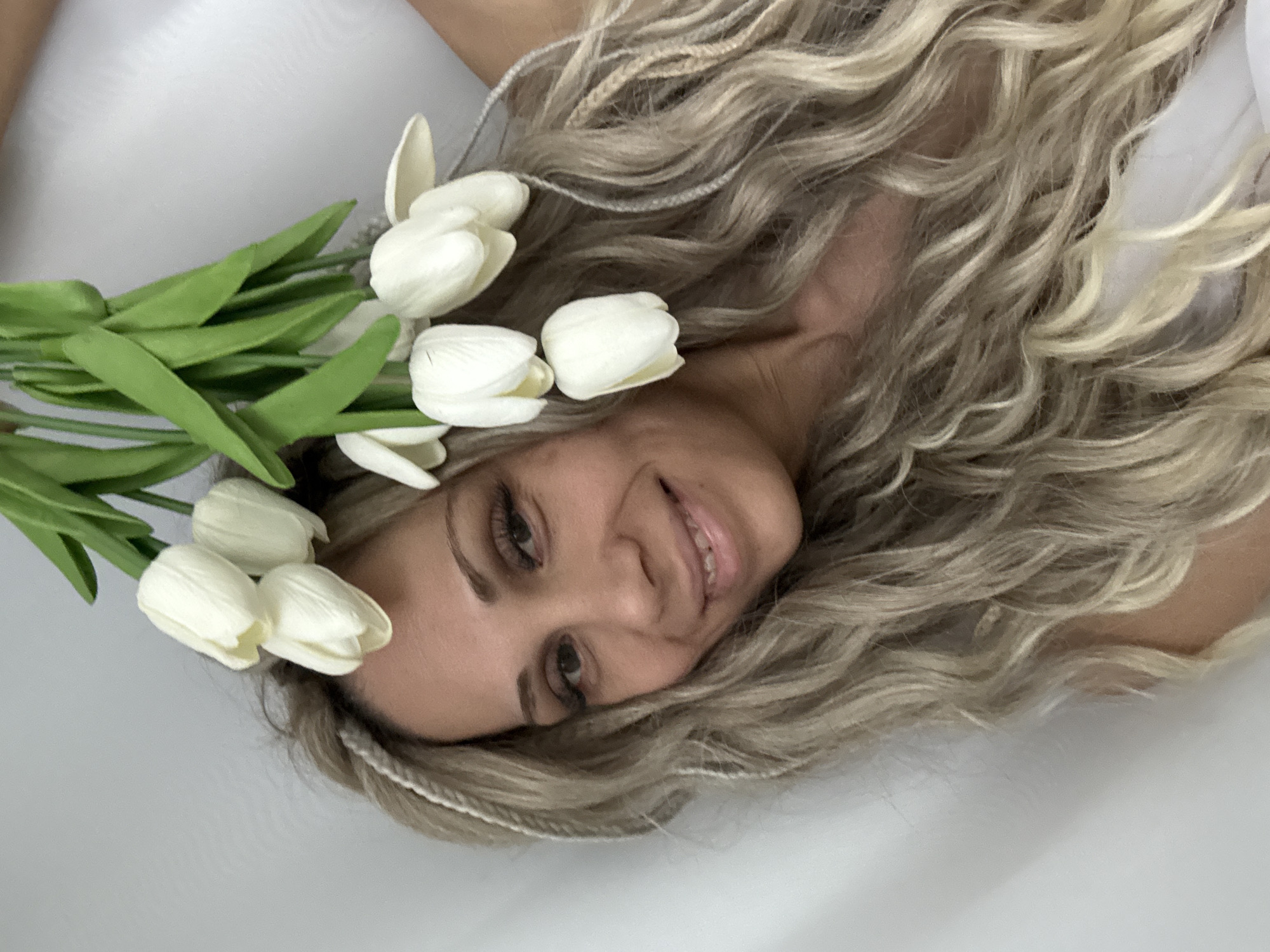 A woman with long, wavy blonde hair is lying on a white surface. She is wearing a crown of white tulips. The photo appears to be taken indoors. There is no verifiable information about the specific location.