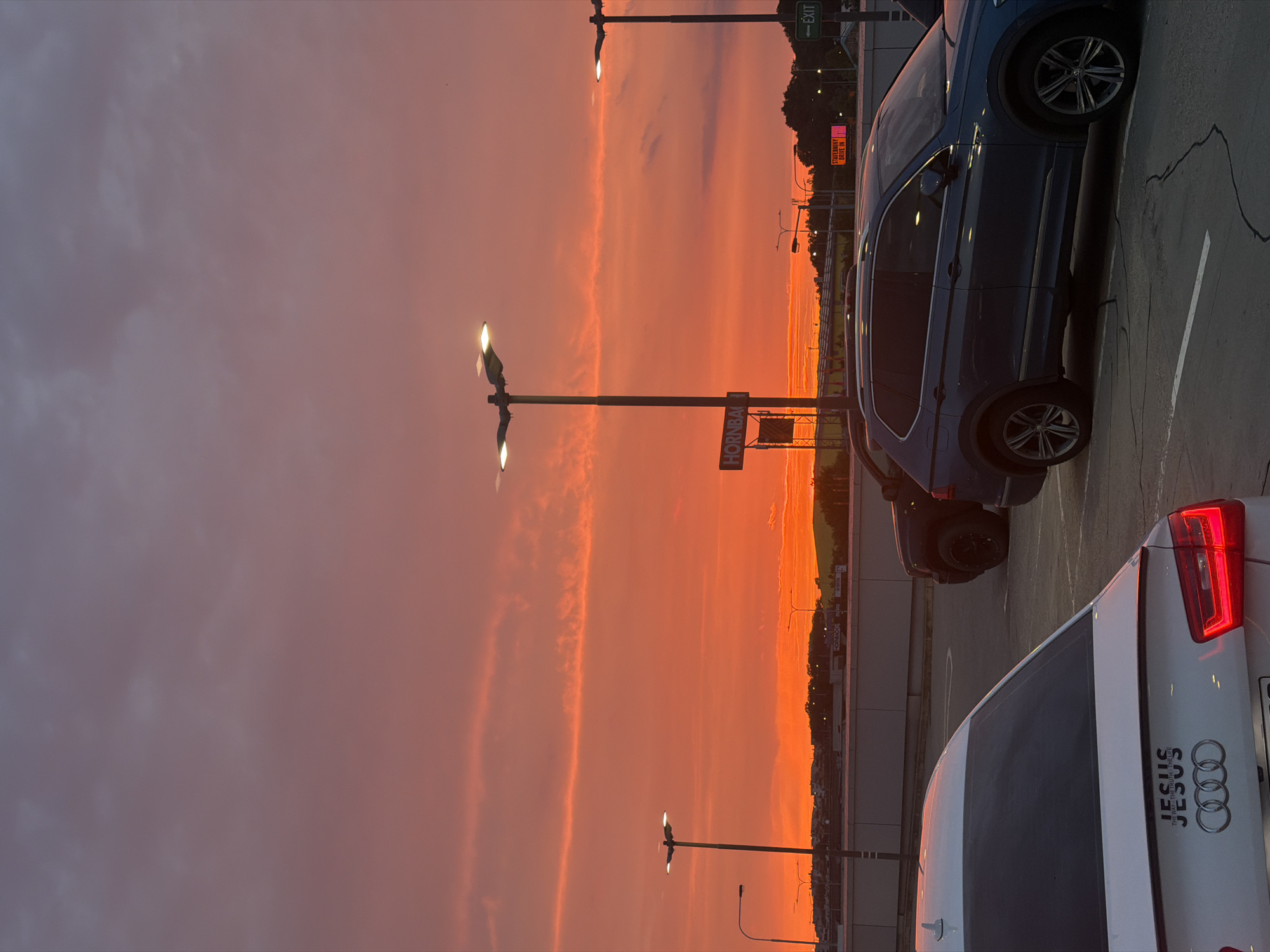 A panoramic, eye-level, outdoor shot captures a vibrant sunset over a city that is likely Prague, Czechia, given the presence of "HORNBACH" signage and the general architecture. The sky is a dramatic gradient of fiery orange, transitioning to soft pink and purple hues, with thin, wispy clouds streaking across the horizon. In the foreground, a white Audi sedan is parked, its rear prominently displaying the "JESUS THE WAY THE TRUTH THE LIFE" decal above the Audi rings. To the right of the Audi, a dark blue Volkswagen SUV is parked. In the middle ground, multiple streetlights with illuminated bulbs stand tall, casting a modern glow against the dusky sky. A "HORNBACH" sign is visible on a building in the distance. Further into the background, a green "EXIT" sign points to the right, suggesting a traffic or parking area. The scene is set on what appears to be a paved parking lot, with visible cracks in the asphalt. The overall atmosphere is serene, with the natural beauty of the sunset dominating the urban landscape.