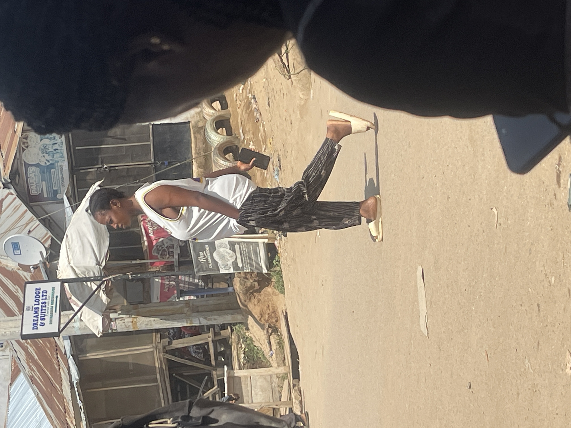 In an outdoor, sunlit scene, a person with dark hair is captured from a high angle, partially obscured by the foreground. They are dressed in a white tank top and black and white striped pants, with white sandals on their feet. The person is holding a black smartphone in their right hand and appears to be walking or standing on a paved surface.
Behind the subject, a street scene unfolds with various buildings and objects. To the left, a white satellite dish is mounted on a corrugated metal roof. A sign for "DREAMS LODGE & SUITES LTD" is visible, with a phone number listed. Further back, several stacks of tires are piled against a wall. There are glimpses of other people and structures, suggesting a busy urban or semi-urban environment. The overall lighting indicates it is daytime, likely sunny. The presence of the satellite dish and the style of the buildings are consistent with a location in Abuja, Nigeria.