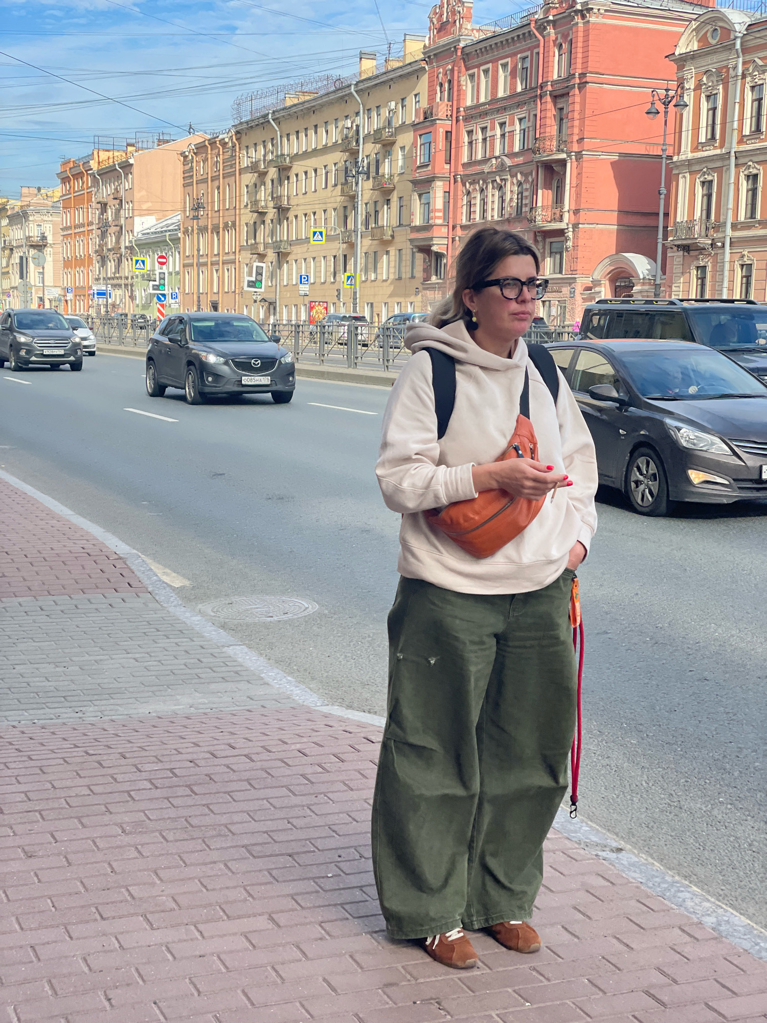 A woman stands on a red brick sidewalk in Saint Petersburg, Russia. She is wearing a light-colored hoodie, dark green wide-leg pants, brown shoes, and glasses. A small orange fanny pack is worn across her chest, and a black backpack is on her shoulders. A red leash hangs from her right side. In the background, a street with several cars and multi-story buildings lines the road. The sky is partially cloudy. The buildings are of various colors including pink, yellow, and beige, and feature balconies and ornate architectural details. Traffic lights and street signs are visible.