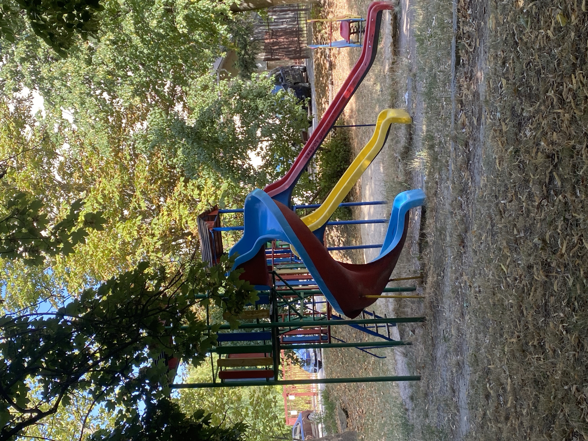 A playground structure with multiple slides is visible in the foreground, set against a backdrop of trees and residential buildings. The playground features a blue slide, a yellow slide, and a red slide, all connected to a multi-level platform. The ground is covered in dry grass and fallen leaves. In the background, a street with parked cars and a fence can be seen. The scene suggests an outdoor urban environment, likely a public park or residential courtyard, in Chișinău, Moldova. The overall condition of the playground appears to be aged, with some wear and tear visible on the slides and surrounding area.