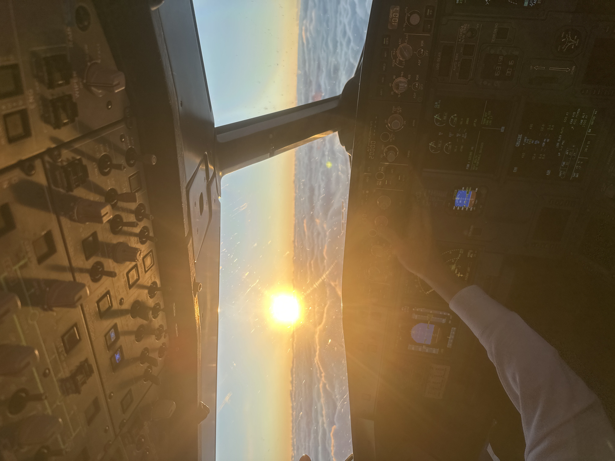 The image depicts the interior of an airplane cockpit, likely a commercial airliner, during what appears to be sunrise or sunset. The sun is visible through the front windows, illuminating the control panels on the left and right sides of the cockpit. A pilot's arm, clad in a white sleeve, is visible on the right side of the frame. The control panels are filled with numerous switches, knobs, and displays. The instruments show flight data, including altitude, speed, and navigational information. The location is not Khimki, Russia; it is impossible to determine the geographical location from this image.