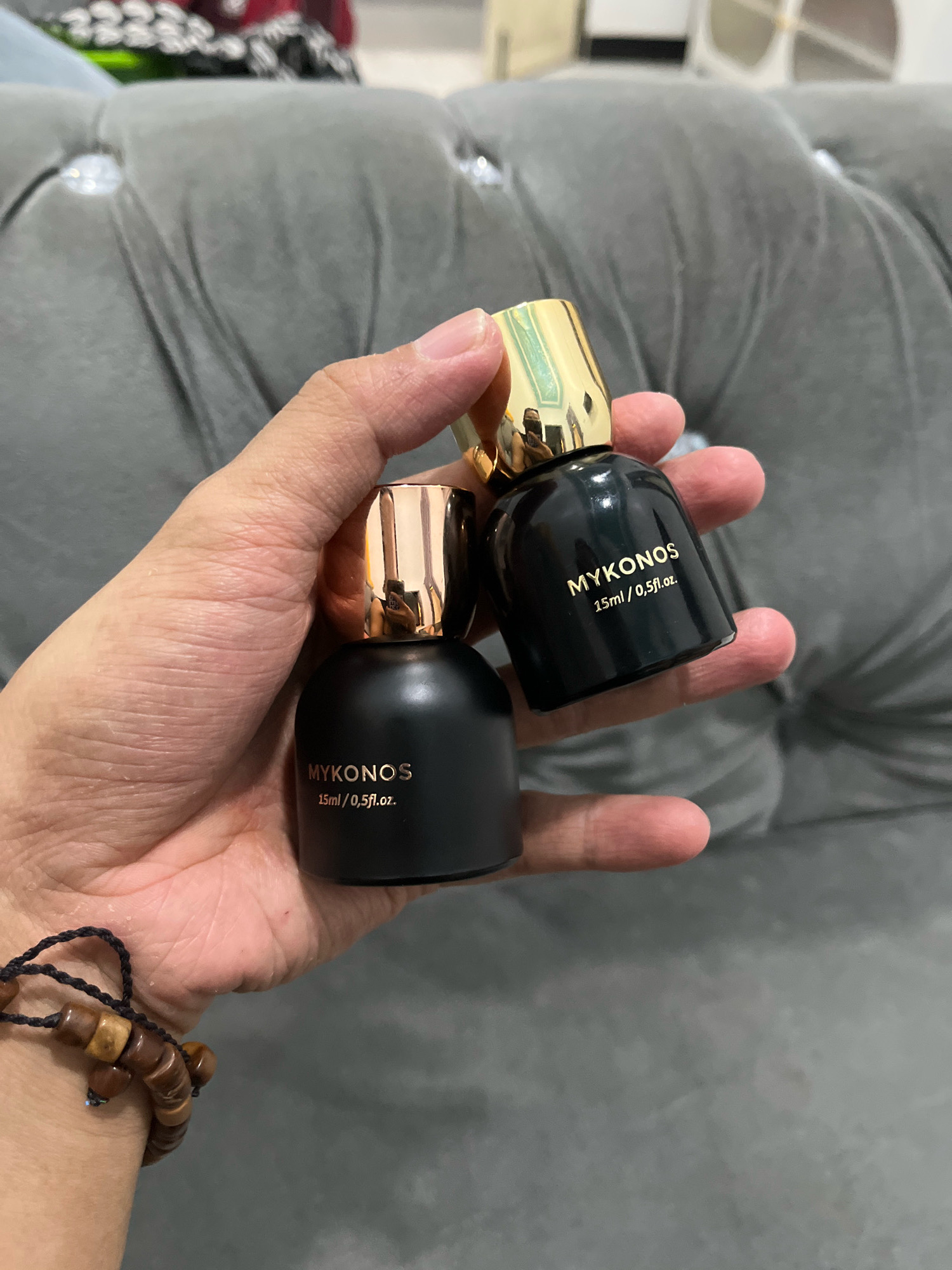 Two small black perfume bottles with gold caps are held in a person's hand. The bottles have "MYKONOS" and "15ml / 0.5fl.oz." printed on them in gold lettering. The background is a tufted gray sofa. The person is wearing a beaded bracelet on their wrist. There is no indication of the location in Tungkaran Pangeran, Indonesia.