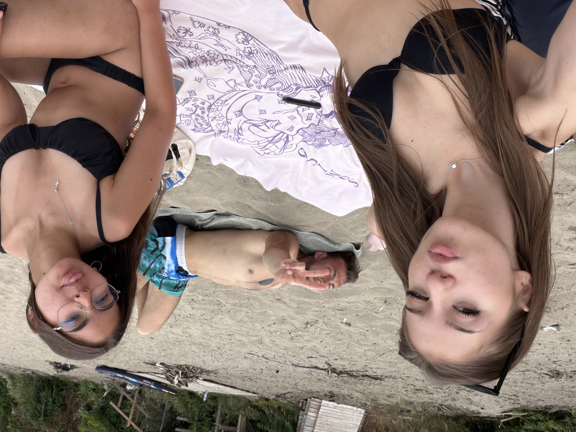 The image shows a beach scene with three people and several objects on the sand. In the foreground, two young women are positioned close to the camera, with one on the left and the other on the right. Both are wearing black bikinis. The woman on the left is looking at the camera with her lips pursed and sunglasses resting on her head. The woman on the right is wearing glasses and has her tongue sticking out slightly. Between the two women is a patterned beach towel. In the background, a man is lying on his back on a towel, holding up two fingers in a peace sign. He is wearing patterned swim shorts. The environment appears to be a sandy beach with some vegetation and wooden structures in the background. A paddleboard is visible on the sand further back, suggesting the location is near water. The scene is consistent with a day at the beach in Tsarevo, Bulgaria, a coastal town known for its beaches and recreational activities.