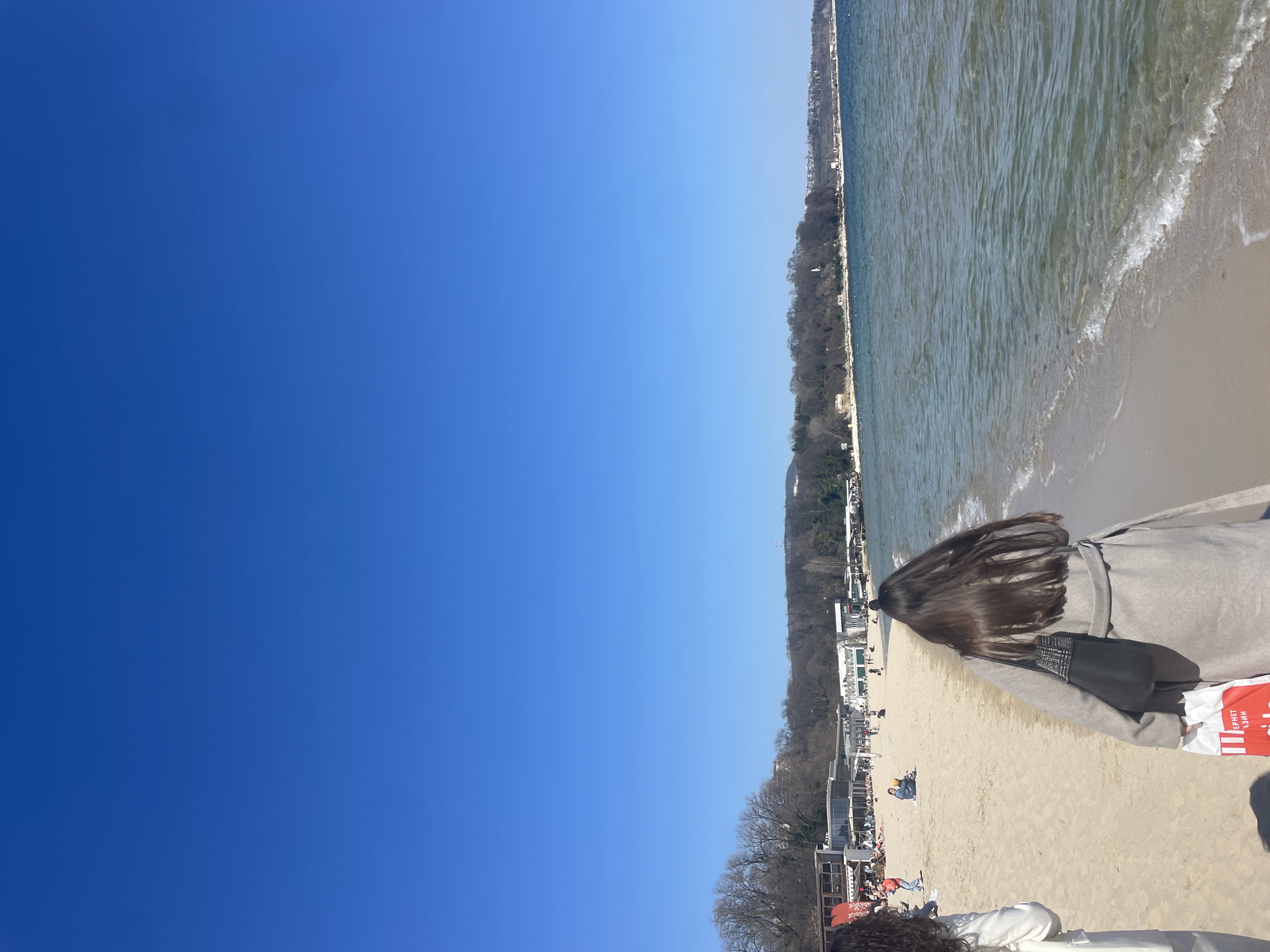 The image depicts a beach in Varna, Bulgaria. A clear blue sky takes up half the image, transitioning to a sandy beach bordering the Black Sea. People are present on the beach, some sunbathing and others walking. Vegetation lines the area between the beach and the buildings. There are buildings along the coastline. Two people, facing away from the camera, are located towards the right of the image, and one is holding a red shopping bag. One has long hair, and wears a light-colored coat.
