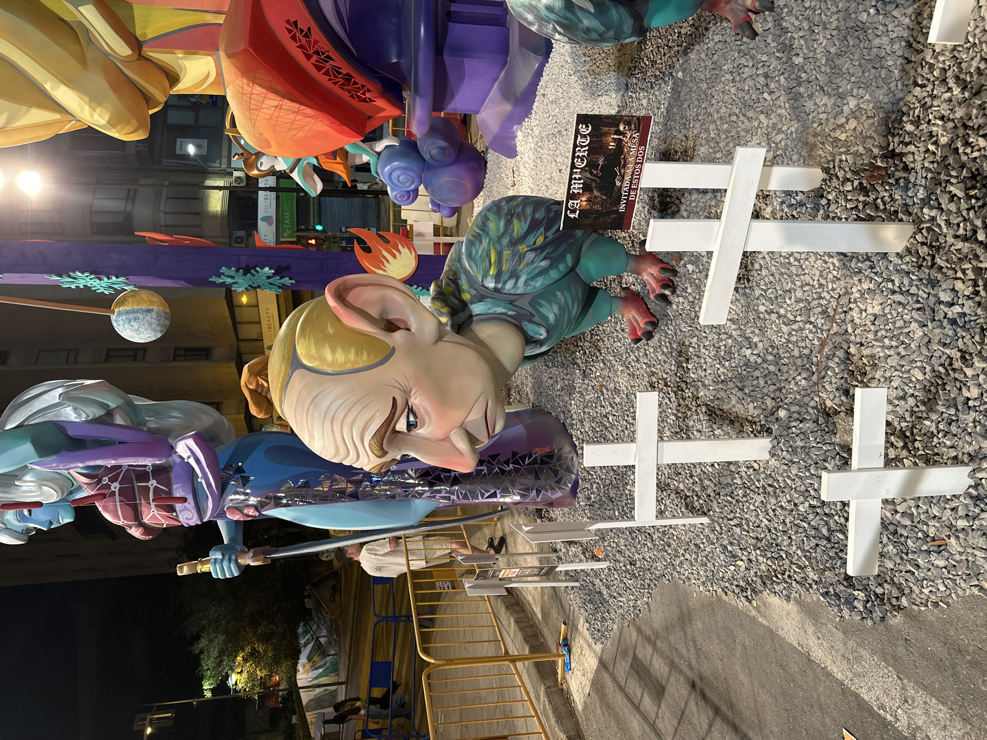 This image captures a nighttime scene of a monument, likely a part of a festival like Las Fallas or a similar local celebration, in Alicante, Spain. The foreground is dominated by a large, exaggerated sculpture of a human head with a weathered face, large ears, and a stern expression. This head is attached to a bird-like body with colorful, feathered patterns and red, clawed feet. In front of this sculpture, several white crosses are planted in a bed of gravel, some appearing as gravestones.
To the left, a towering, stylized humanoid figure with a blue face and silver hair holds a golden sword. This figure is adorned with purple and red elements. Behind these main sculptures, other colorful, abstract figures and architectural elements are visible, illuminated by streetlights and ambient city lighting. A sign with Spanish text, "LA MUERTE INVITADA A LA MESA DE ESTOS DOS," is placed near the bird-like sculpture, implying a theme of death or mortality.
In the background, city buildings and trees are faintly visible, along with yellow safety barriers and what appears to be a street or walkway. A few individuals are present, including one person in the middle ground wearing a light-colored t-shirt and shorts, standing behind the barriers. The overall atmosphere is festive, yet with a somber undertone due to the crosses and the "death" signage. The lighting suggests it is evening or night.