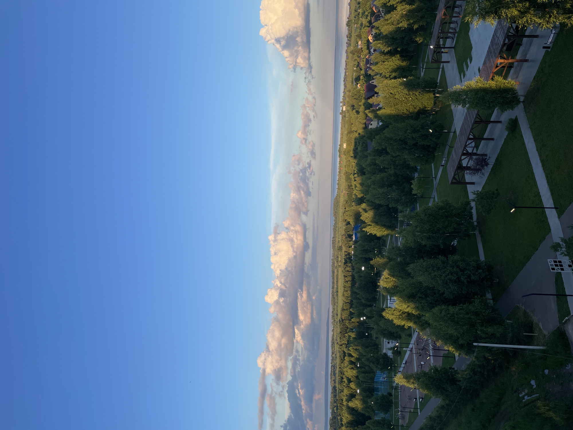 The image is an aerial view of a park area in Laishevo, Russia, at sunset. The sky is a clear blue with scattered, large, fluffy clouds tinged with pink and orange. In the foreground, there are paved pathways winding through a grassy park dotted with numerous tall, leafy green trees. Several open-air wooden structures, resembling pergolas or covered seating areas, are visible. Streetlights are interspersed throughout the park. In the mid-ground, the park transitions into a denser forest, beyond which lies a body of water, likely the Kama River, and a residential area with houses and rooftops. In the far distance, the horizon is marked by a tree line and the expanse of the river or lake. No people are visible in the scene. The overall atmosphere is peaceful and serene, characteristic of an early evening in a suburban or rural setting.