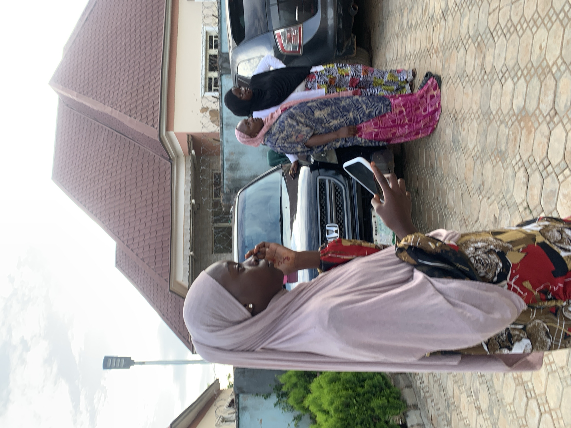 This image captures an outdoor scene in the city of Jos, Nigeria, featuring four individuals, multiple vehicles, and residential buildings under an overcast sky.
In the foreground, a young Black woman is seen in profile, facing slightly right. She wears a light pink/beige hijab covering her head and shoulders, and a patterned garment with red, black, and brown designs. Her right hand is raised to her face, touching near her nose, and exhibits a faint reddish pattern, possibly henna, on her palm. She wears a single stud earring in her right ear. Her left hand holds a white smartphone, partially visible.
In the mid-ground, to the right, three other Black women are standing near two parked dark-colored SUVs. One woman wears a pink hijab and a blue, white, and brown patterned kaftan-style top over a pink patterned skirt. She stands beside a dark Honda SUV. Behind her, another woman wears a black hijab, a white long-sleeved top, and a brightly patterned skirt. Her right hand rests on her hip. A third woman or person is partially visible behind the second SUV.
The vehicles include a dark Honda SUV with its front grille and emblem visible, and a dark SUV (possibly a Ford Edge) parked behind it, showing its rear taillight section. The ground is paved with light brown interlocking concrete blocks arranged in a geometric pattern.
In the background, several single-story buildings with distinctive reddish-brown tiled hip roofs are visible. The roofs feature a layered, stepped design. Walls surrounding the properties are light-colored, with sections topped with razor wire. A tall, modern, rectangular LED street light on a pole stands to the far left against the bright, overcast sky. Green, manicured shrubs are visible on the left side of the frame. The scene is brightly lit by natural daylight.