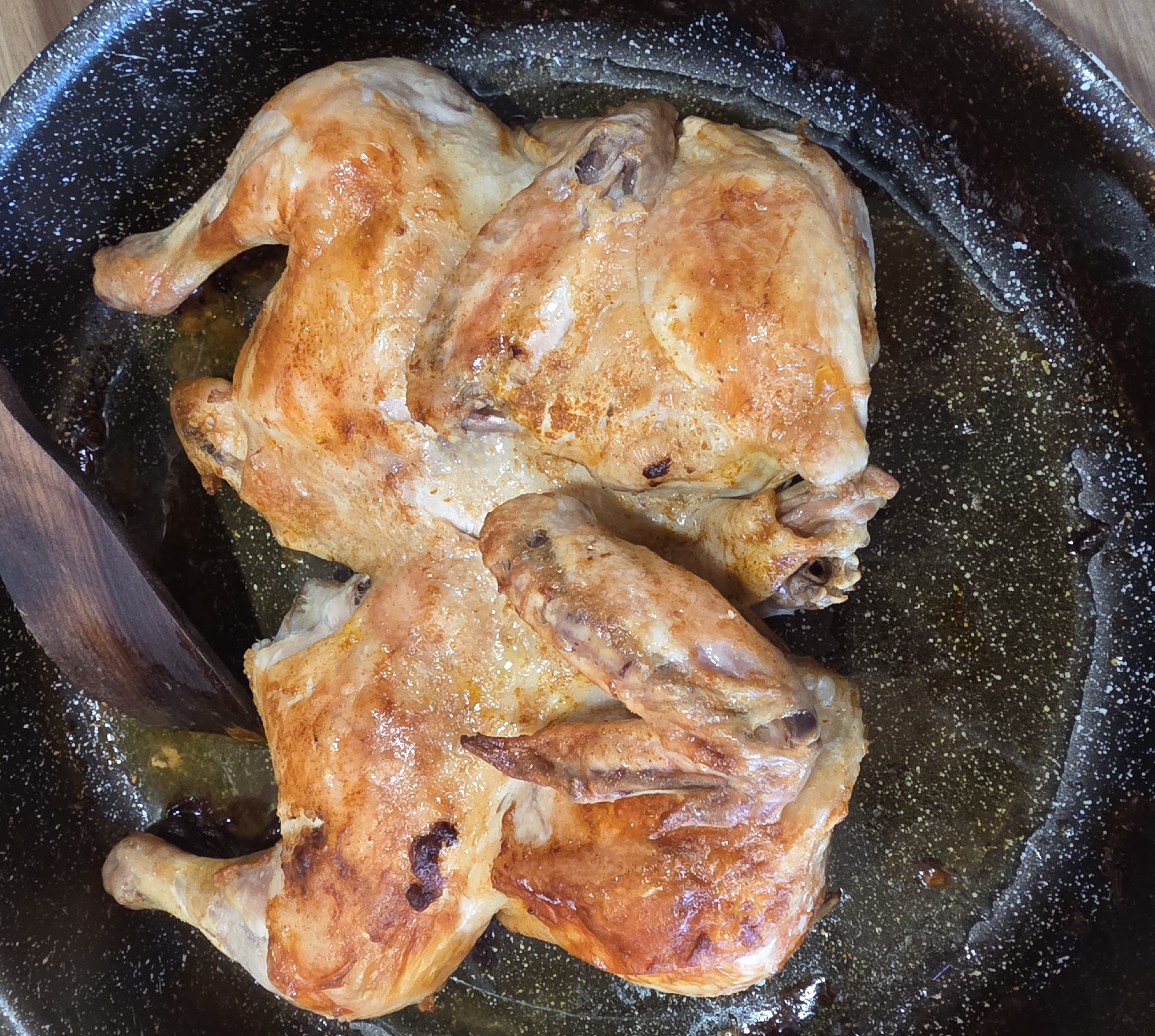 The image shows a roasted chicken cut in half and placed in a black frying pan with some cooking oil. A wooden spoon lies near the chicken. The scene appears to be indoors, on a wooden surface. The location in Novachene, Bulgaria is not discernible from the image.