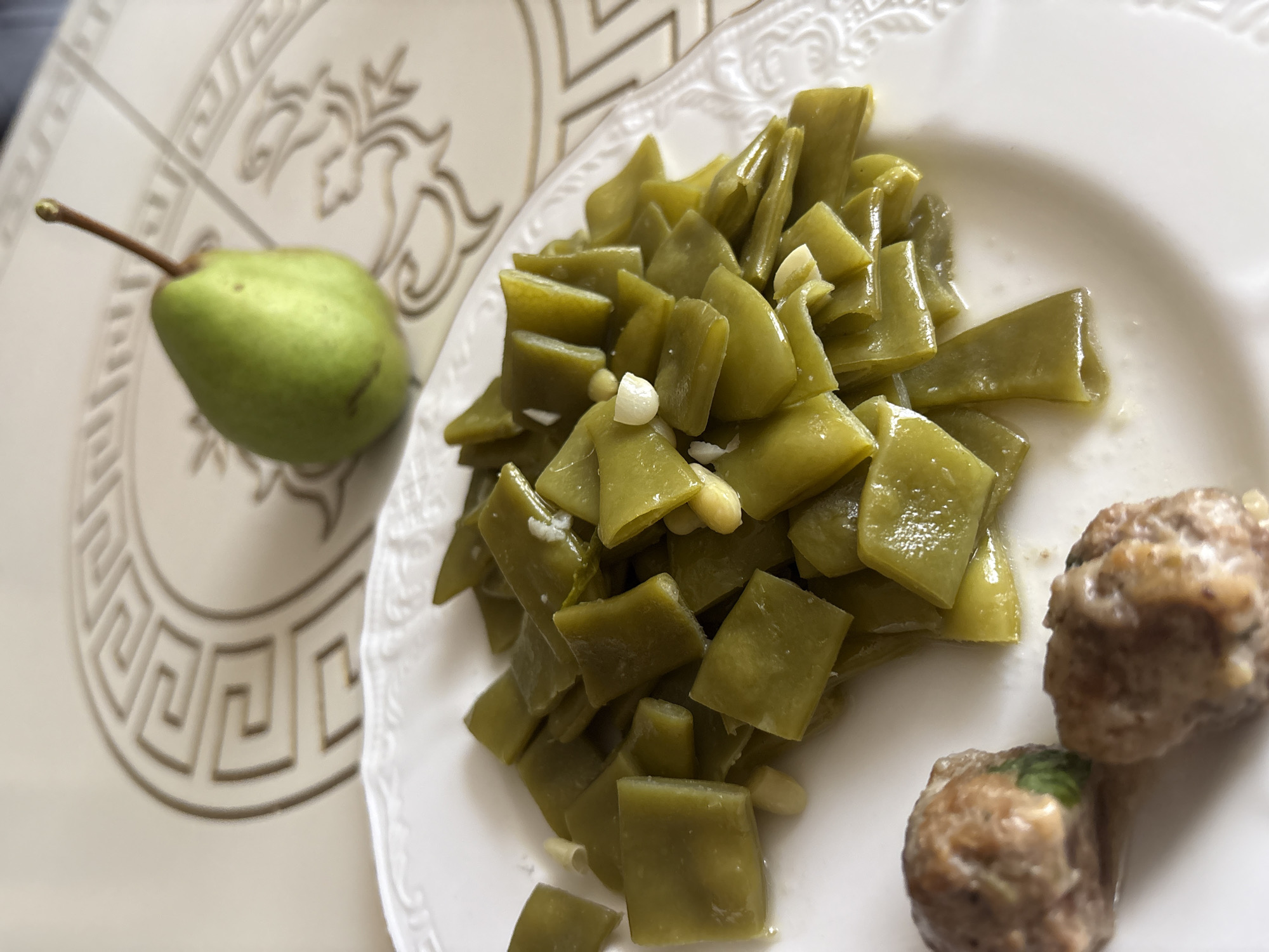 A white plate with a decorative rim sits on a table with a patterned surface. The plate contains a portion of green beans, some of which appear to be cooked with small pieces of garlic. Two small, round meatballs are visible on the left side of the plate. In the background, to the right of the plate, a single green pear with a thin stem rests on the table. The table surface has a decorative circular motif with Greek key patterns. There are no people visible in the image. The location is Georgiyevsk, Russia.