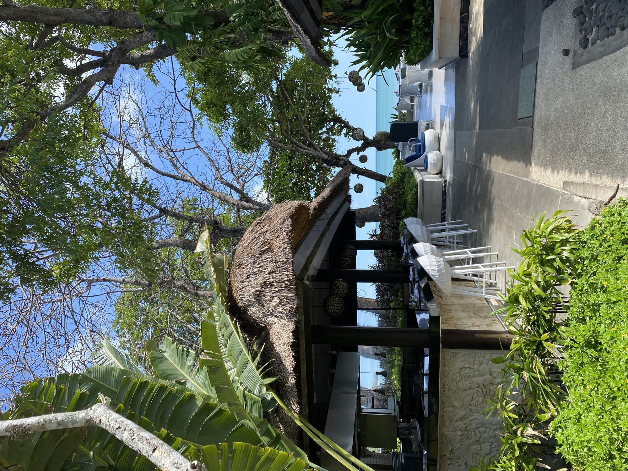 The image depicts an outdoor setting in Kuta Selatan, Indonesia. The scene features a thatched-roof structure surrounded by lush tropical vegetation, including palm trees and other greenery. Beyond the structure, the blue ocean and horizon are visible. A tiled area with outdoor seating, including white chairs and blue cushions, suggests a dining or lounge space. A carved stone wall is adjacent to the seating area. The sky is clear and blue with scattered clouds.