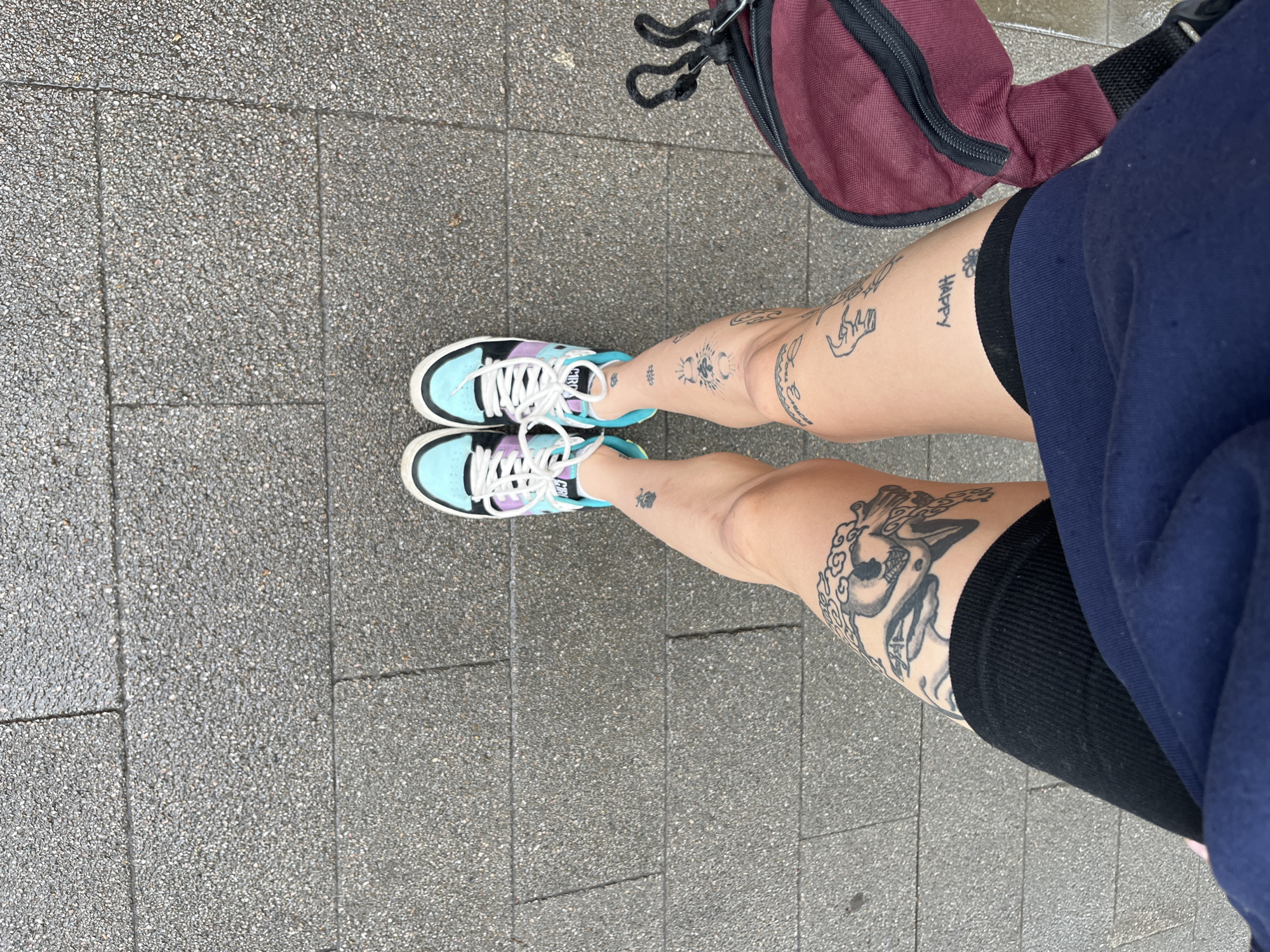The image shows a person's legs from the knees down, standing on a gray, pebbled sidewalk. The person is wearing black shorts, a navy blue sweatshirt, and colorful sneakers with white laces. There are several tattoos on the person's legs. To the left of the person's legs is a burgundy-colored bag with a black strap. The sidewalk appears to be wet, suggesting recent rain or damp conditions. The scene is outdoors in an urban environment. There is no information in the image to indicate the location is Krasnoyarsk, Russia.