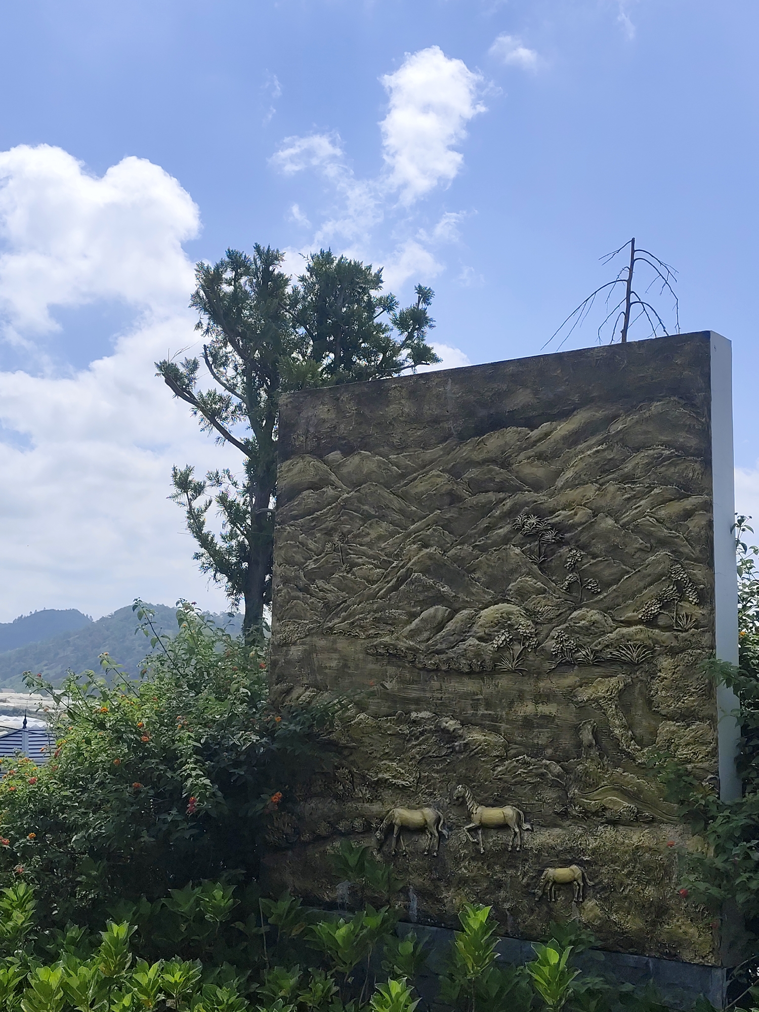 This outdoor vertical shot captures a large, rectangular sculptural wall or panel positioned within a verdant landscape under a bright, cloudy sky in Lạc Dương, Vietnam. The wall features an elaborate embossed relief depicting a natural scene. The artwork on the wall portrays rugged mountains and hills, interspersed with stylized trees and what appears to be a river or stream winding through the landscape. At the bottom left of the relief, three horses are depicted in various stages of grazing: two larger horses stand side-by-side, and a smaller foal is positioned slightly below and to their right.
In the foreground and around the base of the sculptural wall, lush green bushes and foliage are prominent, some with small orange blossoms. Behind the wall on the left, a tall, vibrant green tree with dense foliage stands tall, its upper branches reaching towards the sky. On the opposite side, above the right edge of the wall, a bare, skeletal tree with thin, leafless branches is silhouetted against the sky. Distant, hazy green mountains or hills are visible in the background on the left, beneath the bright blue sky adorned with puffy white clouds. The scene is bathed in clear daylight, suggesting a sunny morning or afternoon. There are no people visible, and no discernible text. The overall impression is one of natural beauty combined with artistic representation, hinting at a serene and picturesque location.