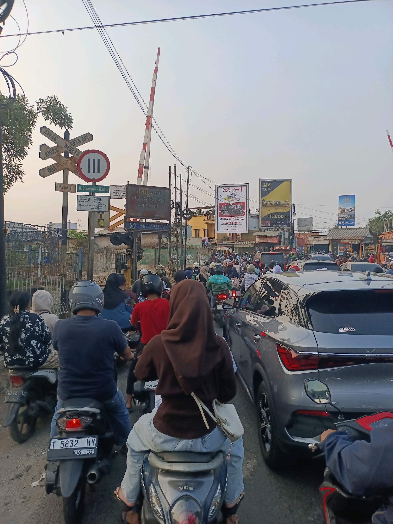 The image captures a busy street scene in Karawang, Indonesia, featuring a significant traffic jam. The foreground is dominated by motorcycles and cars, with people on motorcycles wearing helmets and casual clothing. A gray SUV is prominently visible in the right lane. In the background, a railway crossing gate with red and white stripes is raised, indicating that a train is not currently passing. Several signs are present, including directional signs and traffic warnings like "AWAS KERETA API" (Beware of Train). Billboards and advertisements for various products and services are also visible on the buildings lining the street. The overall environment suggests a typical urban setting with moderate daylight and a clear sky. The traffic is heavy, with vehicles lined up as far as the eye can see.