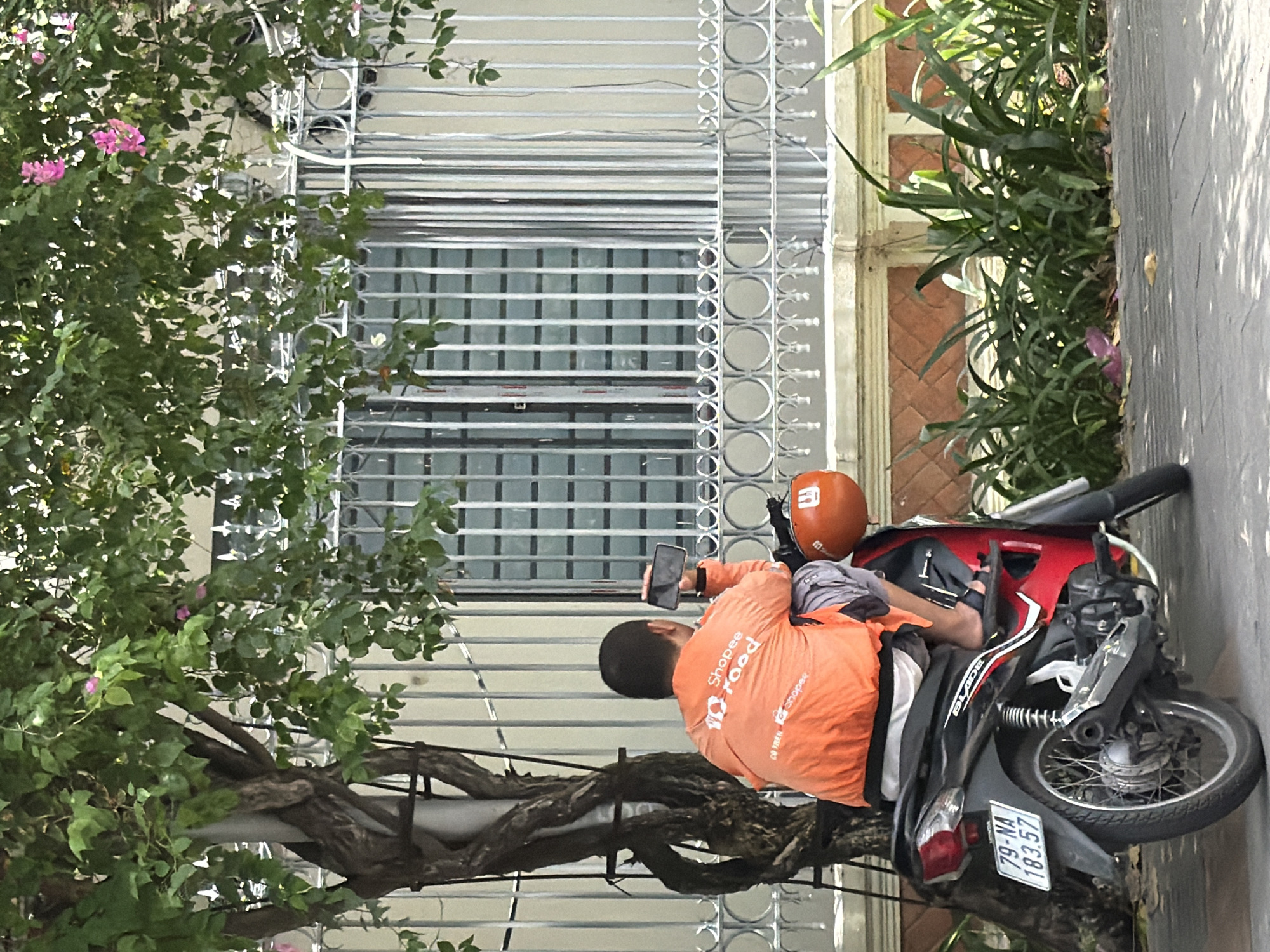 A person, wearing an orange "Shopee Food" jacket and shorts, sits on a red and black motorcycle on a paved street in front of a building. The person is holding a smartphone in their right hand, possibly taking a photo or video. The motorcycle has a license plate that reads "79-NA 183.57". An orange helmet is attached to the back of the motorcycle. A tree with green leaves and some pink flowers is to the left of the person. The building has a white facade with a metal fence and barred windows. Green plants are in front of the building. The image appears to be taken in Nha Trang, Vietnam, given the license plate format which typically starts with the province code (79 for Khanh Hoa province, which includes Nha Trang).