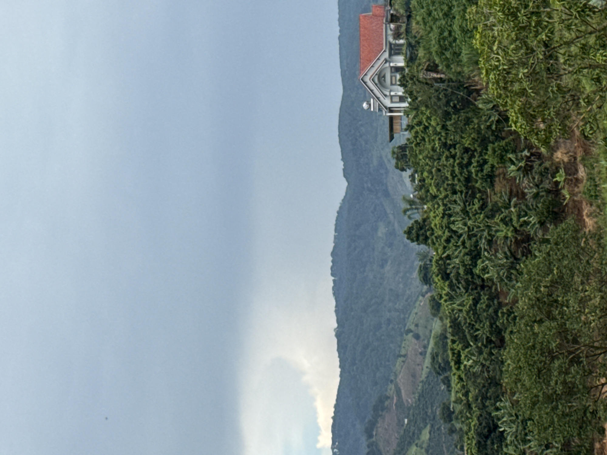 A distant, white, two-story house with a red roof sits on a green, tree-covered hillside under a cloudy, grey sky. The house is the only man-made structure visible in the image. The landscape appears to be rural, with dense vegetation covering the rolling hills. There are no people or animals present in the image. The image depicts a peaceful, natural environment.