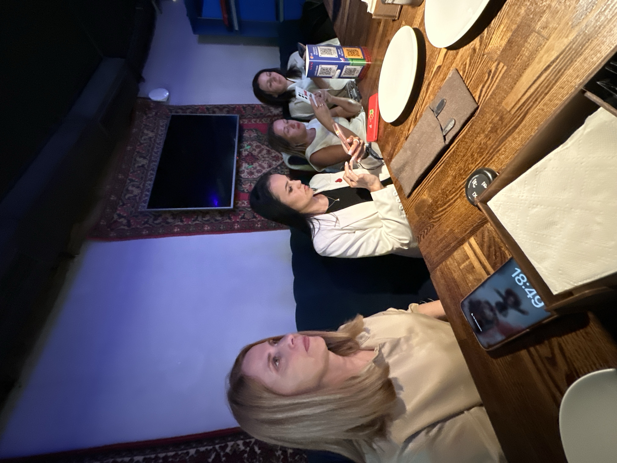 Here is a detailed description of the image:
**Location:** The scene appears to be inside a restaurant or cafe, possibly in Moscow, Russia. This is inferred from the presence of Cyrillic text and the general decor style.
**People:** The image features a group of four women sitting around a wooden table. They are oriented at various angles, suggesting casual conversation and interaction. Each woman has varying poses, clothing, and activities. One is holding a phone with a display visible.
**Objects:** The table is set with plates, napkins, and what appears to be cutlery wrapped in a brown fabric holder. A smartphone rests on the table, displaying the time 18:49 (6:49 PM). In the background is a television and some items on a wall-mounted shelf.
**Actions:** The women seem to be engaged in a social gathering. They are using their phones.
**Environment:** The room has a dim, ambient lighting. A Persian-style carpet is visible along the wall and near the television. The general ambiance suggests a relaxed and informal setting.