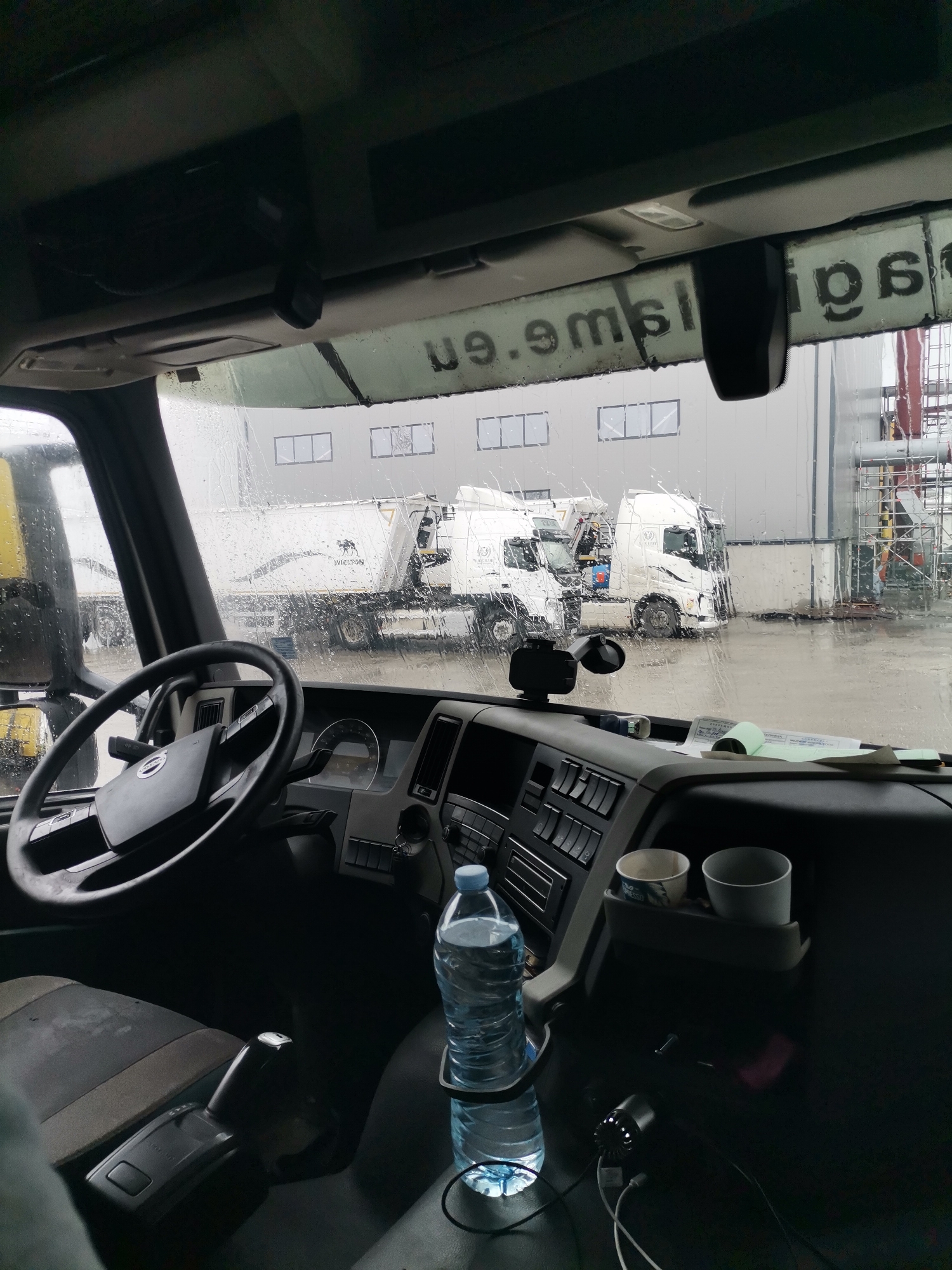 The image provides a view from inside a truck cab looking out through a rain-splattered windshield at an industrial or storage area in Karapelit, Bulgaria. The weather is overcast, and rain is evident from the numerous water droplets on the glass and the wet ground outside.
Outside, two large white trucks, likely dump trucks or cement mixers, are parked side-by-side. Both trucks feature a logo on their trailer bodies, depicting a globe with the text "VISON" or similar beneath it. Behind the trucks stands a long, grey industrial building with several rectangular windows. To the far right, a complex of industrial pipes and scaffolding structures is visible.
Inside the cab, the dark grey dashboard and a black steering wheel, which appears to have a manufacturer's logo (possibly Volvo), are prominent in the foreground. A gear shifter is located to the driver's right. On the passenger side of the dashboard, a small compartment holds two paper cups, one white and one with a design. Below this, a blue plastic water bottle is secured in a holder, with various charging cables connected nearby. Papers, including a green document that resembles a delivery note or logbook, are placed on the dashboard. Overhead, reflected on the top edge of the windshield, inverted text such as "9.9msl.e.9" and "pqs" is visible, which is difficult to fully decipher.
The scene depicts a daytime setting, likely during a work break or a waiting period, amidst rainy conditions at an industrial facility.